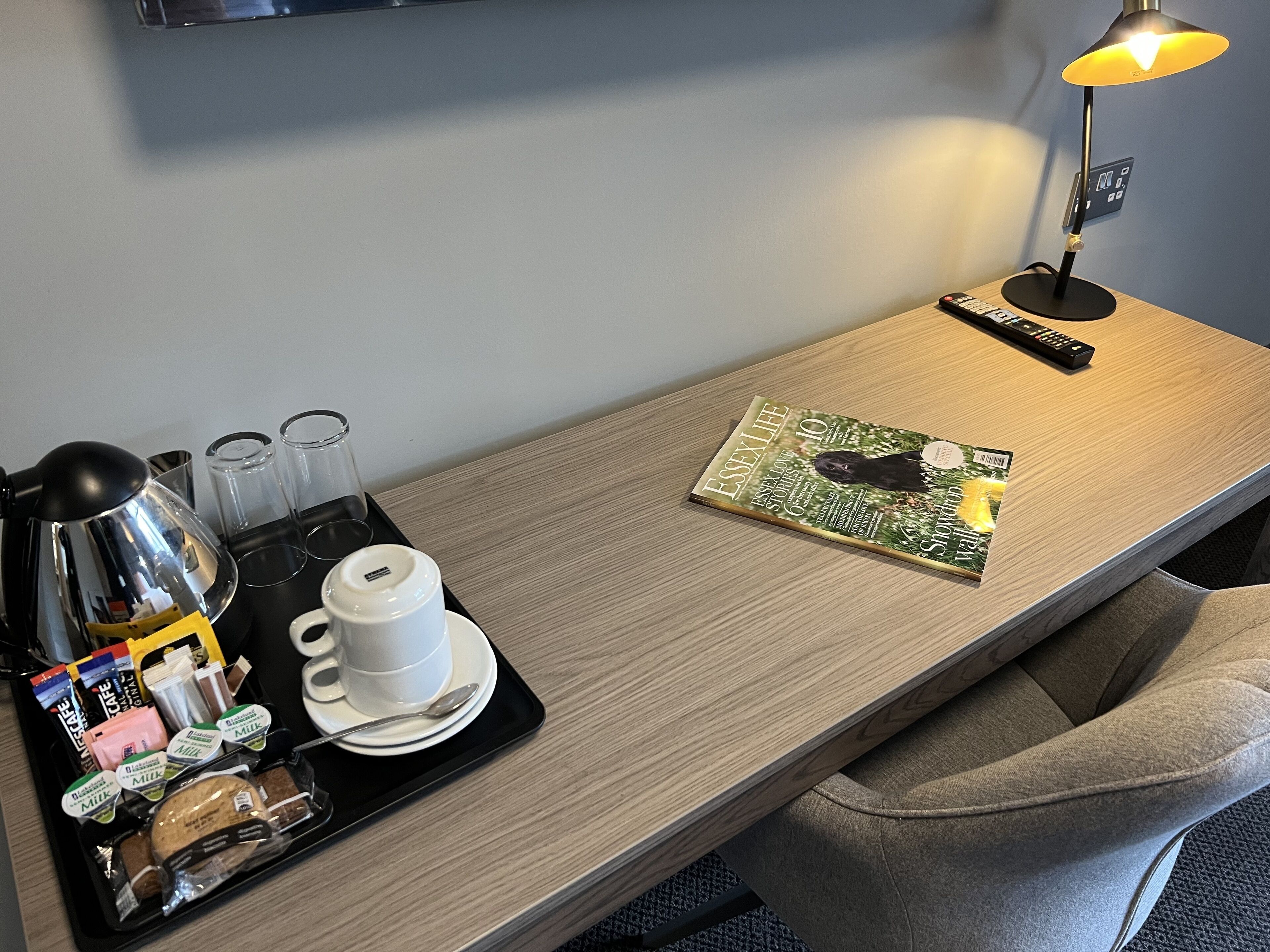 A hotel room with a wooden desk, a magazine, a remote control, a tray with breakfast items, and a coffee maker.