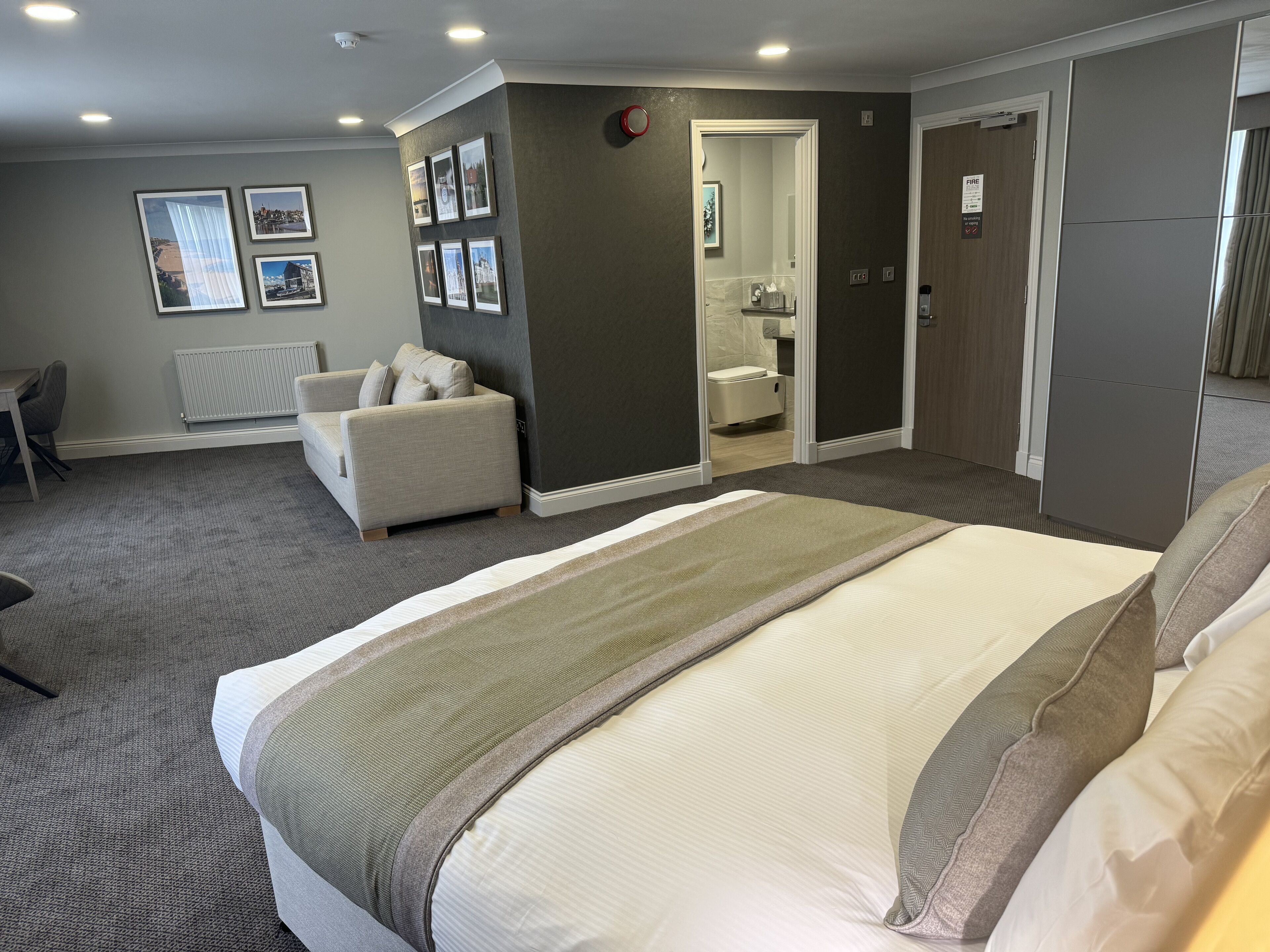 A hotel room with a bed, a sofa, a chair, a radiator, and framed pictures on the wall.