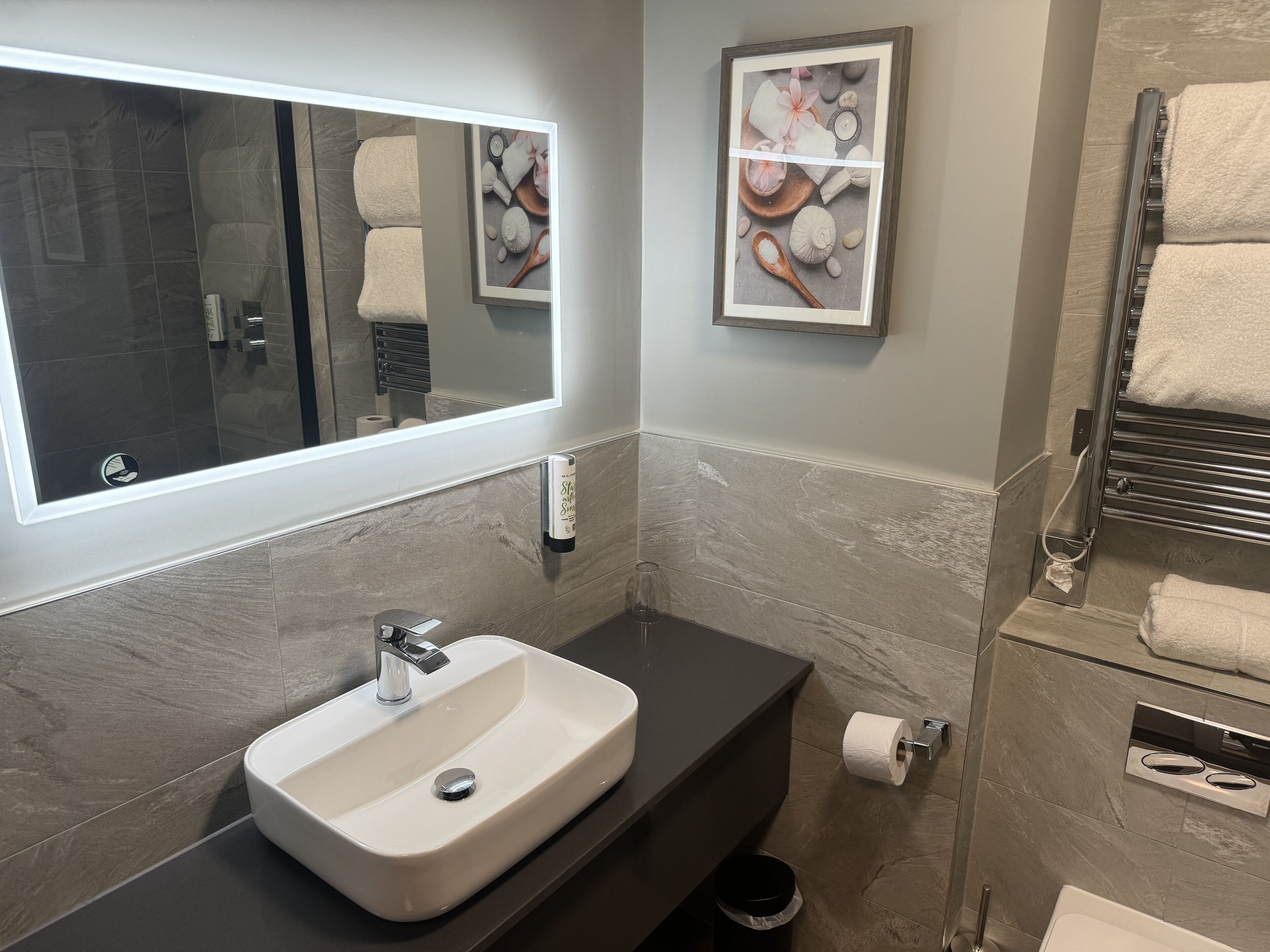 A modern bathroom with a large mirror, a white sink, a towel rack, and a framed artwork on the wall.
