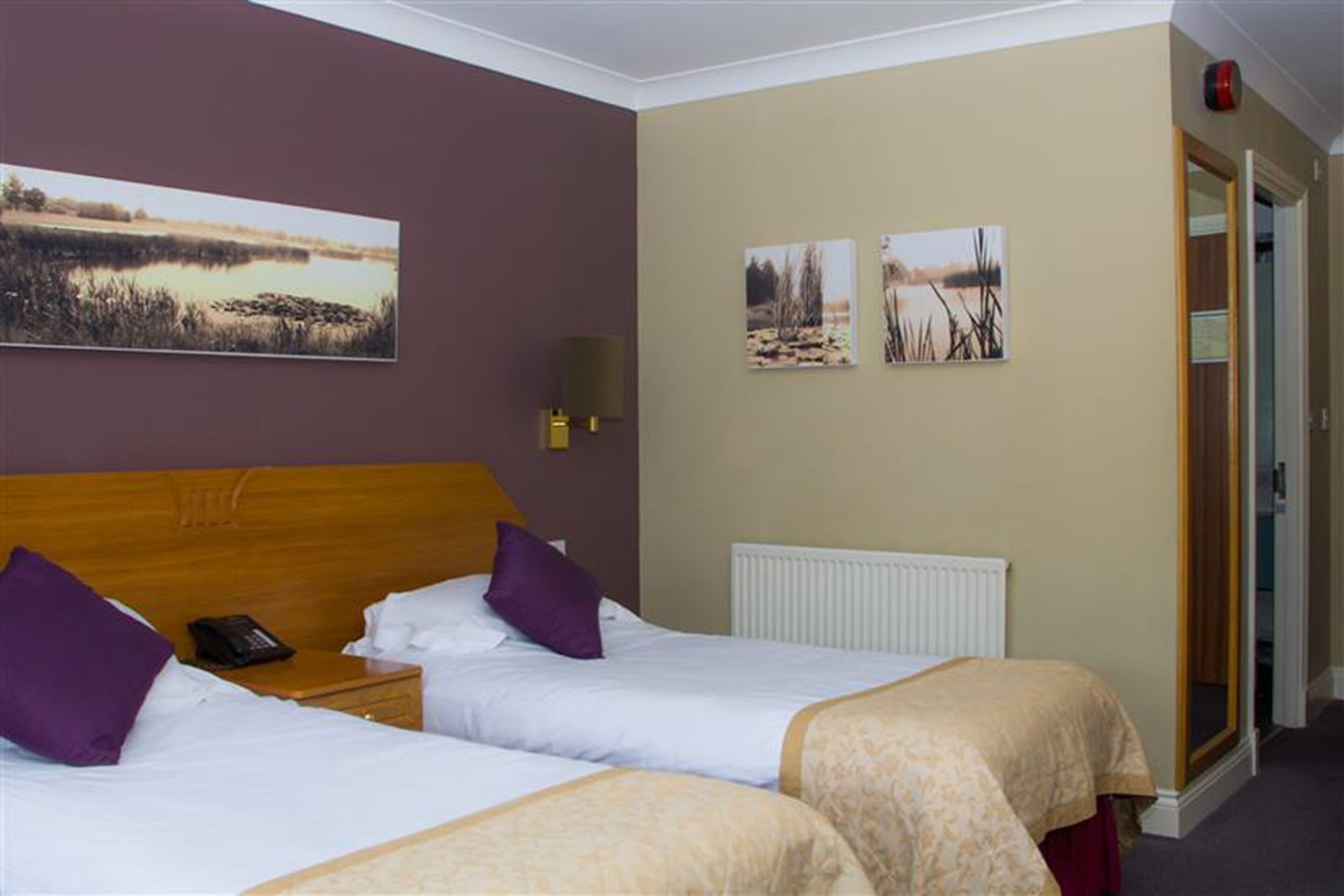 Une chambre d’hôtel avec deux lits simples, une tête de lit en bois, une table de chevet avec un téléphone et des œuvres d’art accrochées au mur représentant des scènes de la nature.