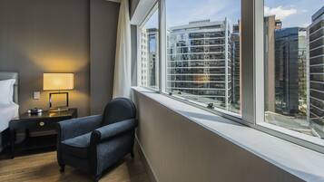 Habitación de hotel con un ventanal grande, una cama, una silla, una mesita de noche con lámpara y vistas a edificios de la ciudad.