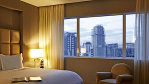 Habitación de hotel con una cama grande, una silla, una mesita de noche con lámpara y vistas a un paisaje urbano a través de un ventanal.