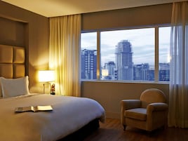 Habitación de hotel con una cama grande, una silla, una mesita de noche con lámpara y vistas a un paisaje urbano a través de un ventanal.