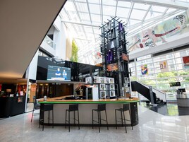 Zona de bar moderna con techo alto y paredes de vidrio, que incluye una barra central con taburetes y una pantalla digital que muestra contenido.