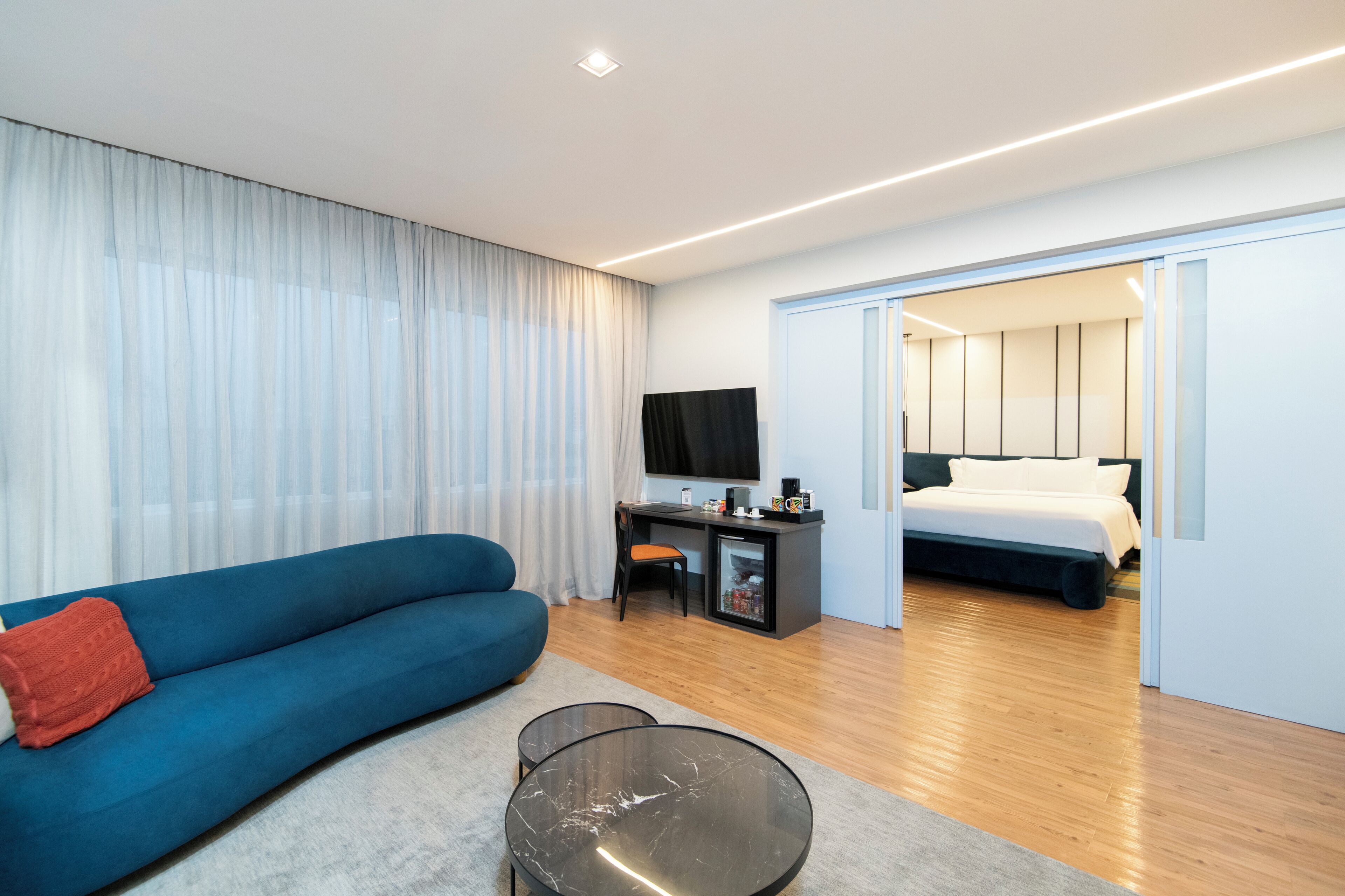 A modern hotel room with a blue sofa, a small desk with a TV, and a separate bedroom with a bed.