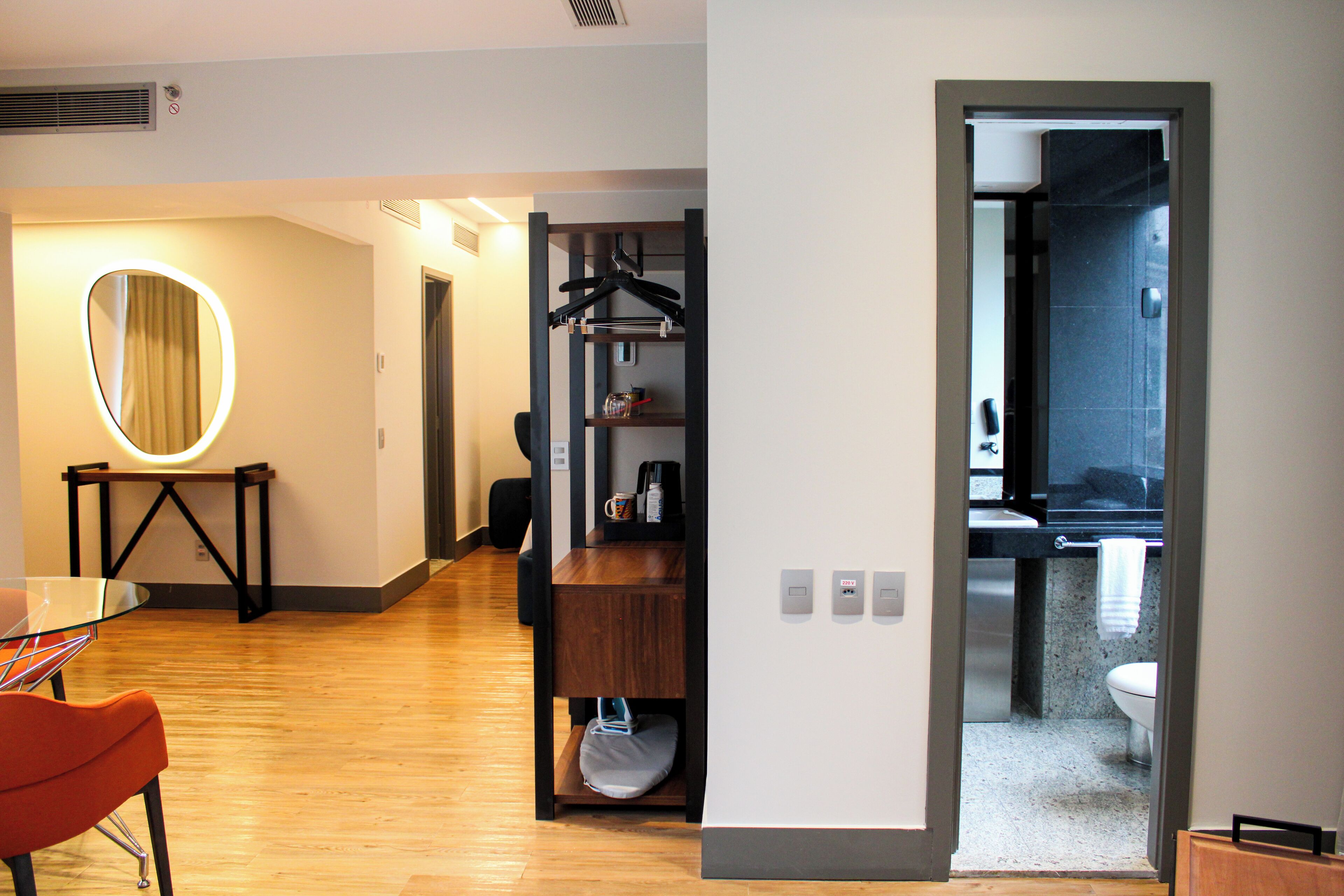 Une chambre d’hôtel avec un plancher en bois, un miroir rond, une table avec des chaises, une étagère avec des vêtements et une vue sur la salle de bain.