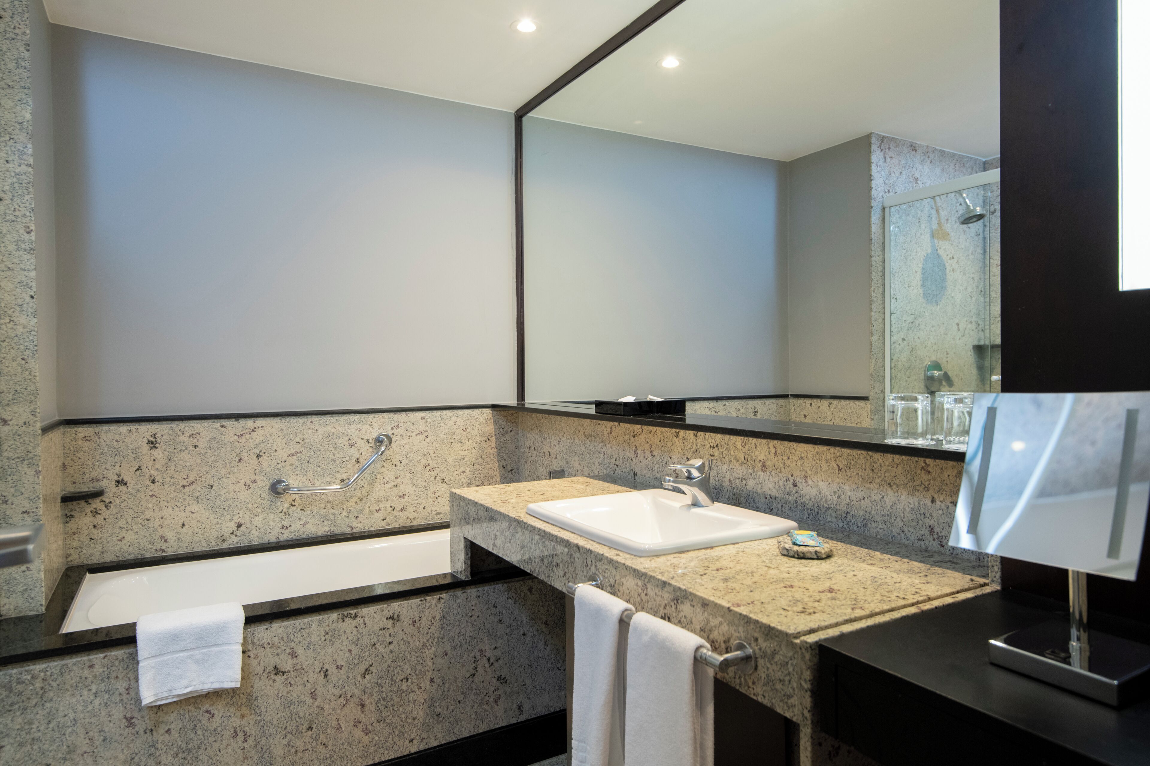 Un baño moderno con encimera de granito, un lavabo blanco, un espejo grande y una ducha con mampara de cristal.