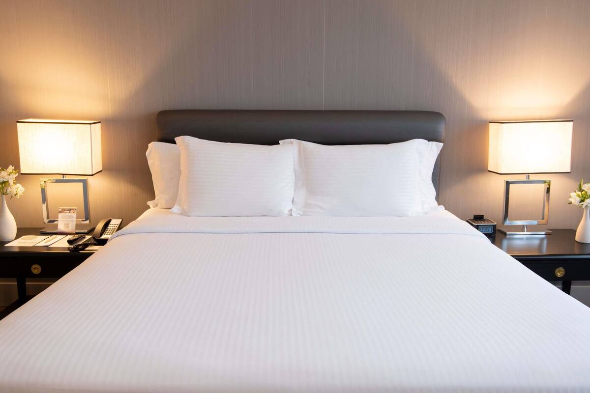 Quarto de hotel com uma cama grande, duas luminárias de cabeceira, mesa de cabeceira com telefone e um vaso com flores.