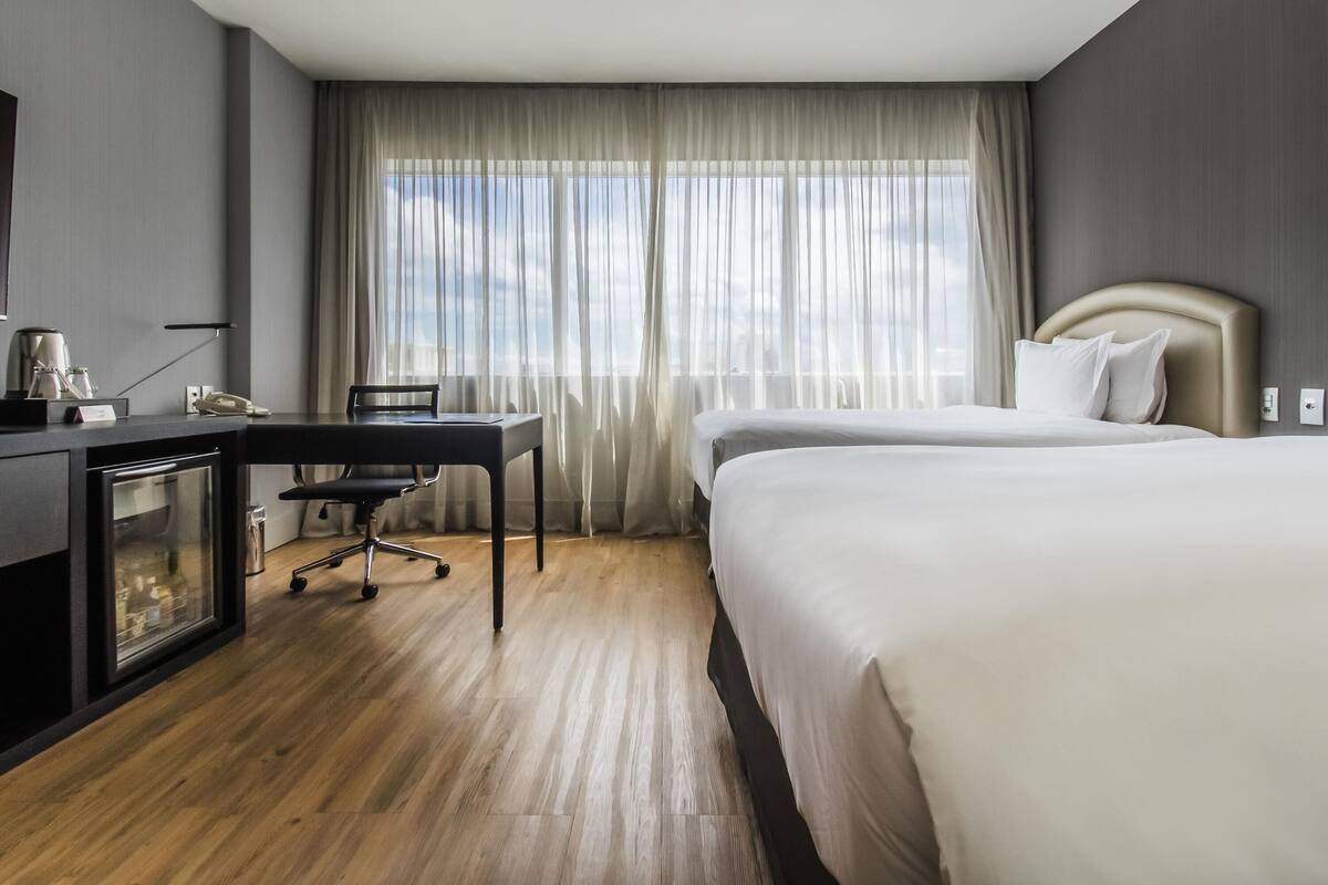 Quarto de hotel com cama, escrivaninha com cadeira, telefone e janela com cortinas.
