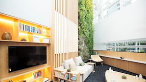 Una habitación moderna y bien iluminada con un televisor de pantalla plana grande, estanterías de madera, un sofá blanco con cojines, una mesita con objetos decorativos y una pared jardín vertical.
