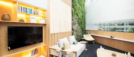Una habitación moderna y bien iluminada con un televisor de pantalla plana grande, estanterías de madera, un sofá blanco con cojines, una mesita con objetos decorativos y una pared jardín vertical.