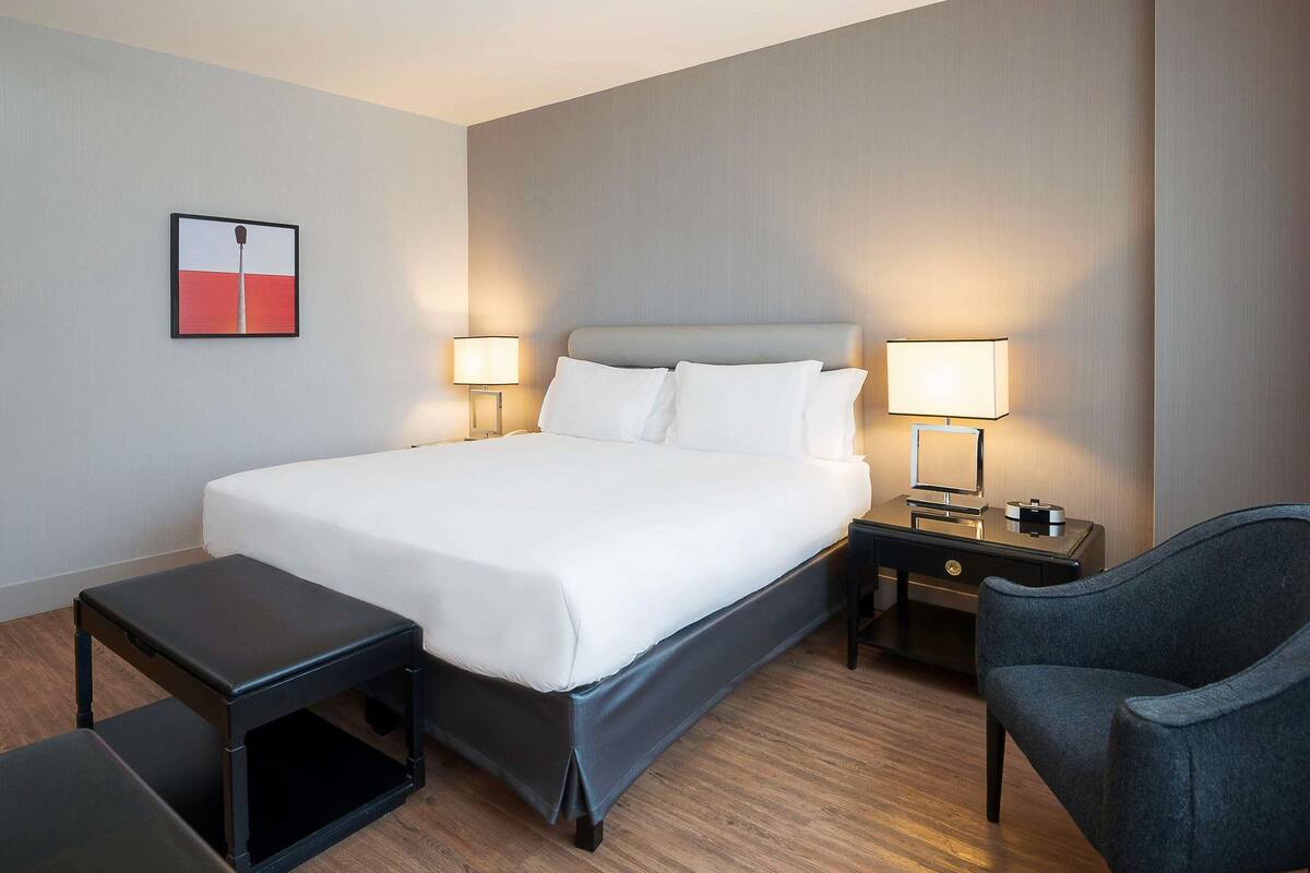 Quarto de hotel com uma cama grande, duas mesas de cabeceira com luminárias, uma cadeira e uma pequena mesa.