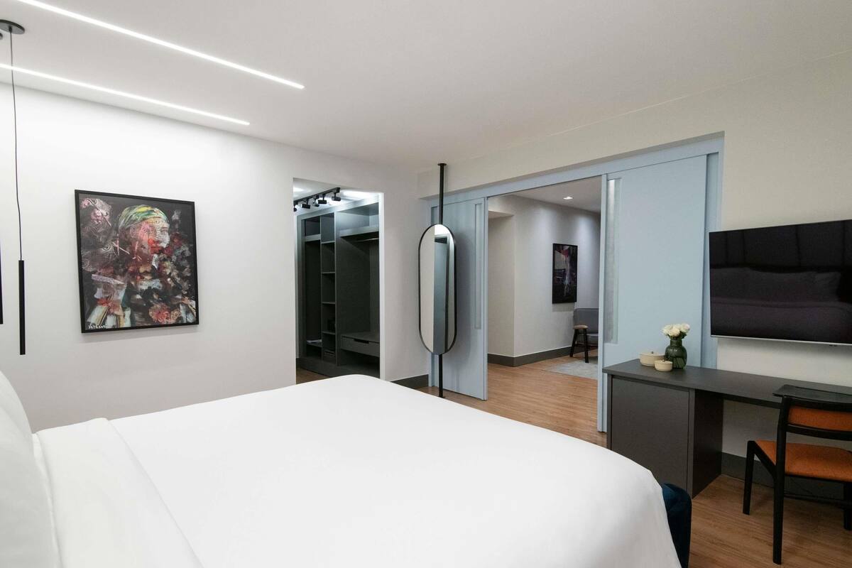 A modern hotel room with a bed, a desk, a TV, a wardrobe, and a painting on the wall.