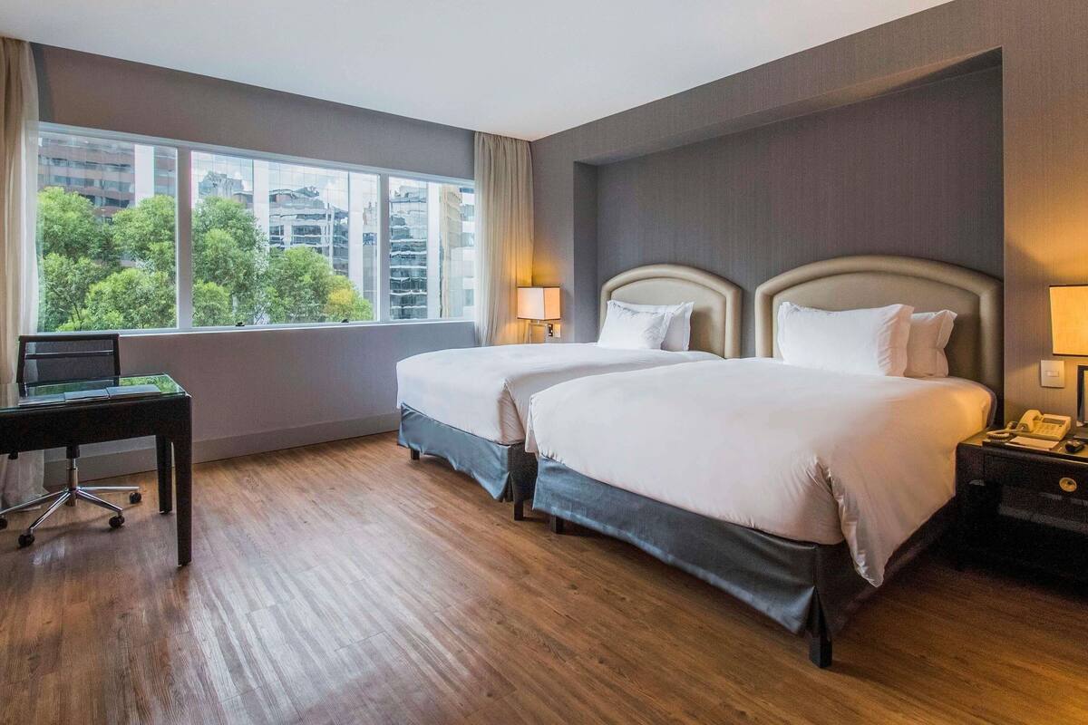 Quarto de hotel com duas camas, uma escrivaninha e uma janela com vista para a cidade.
