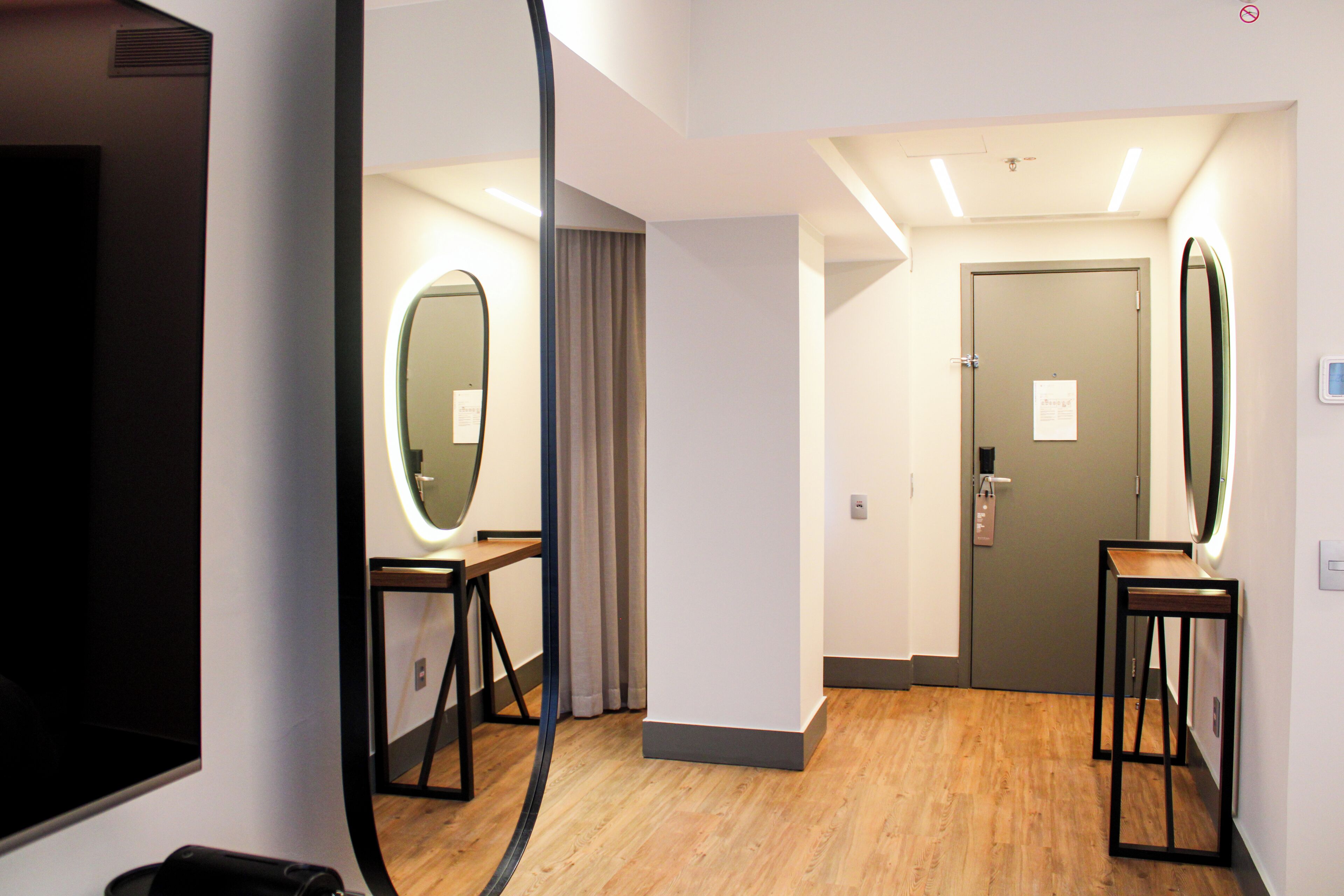 A hotel room with a wooden floor, a mirror, a door, and a lighted wall.