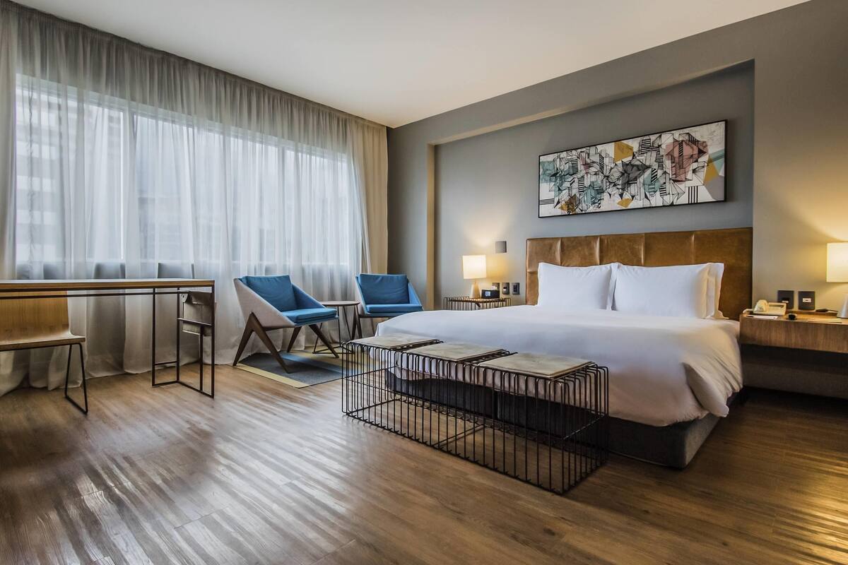 Quarto de hotel com uma cama, duas cadeiras, uma escrivaninha, uma mesa de centro e um quadro na parede.