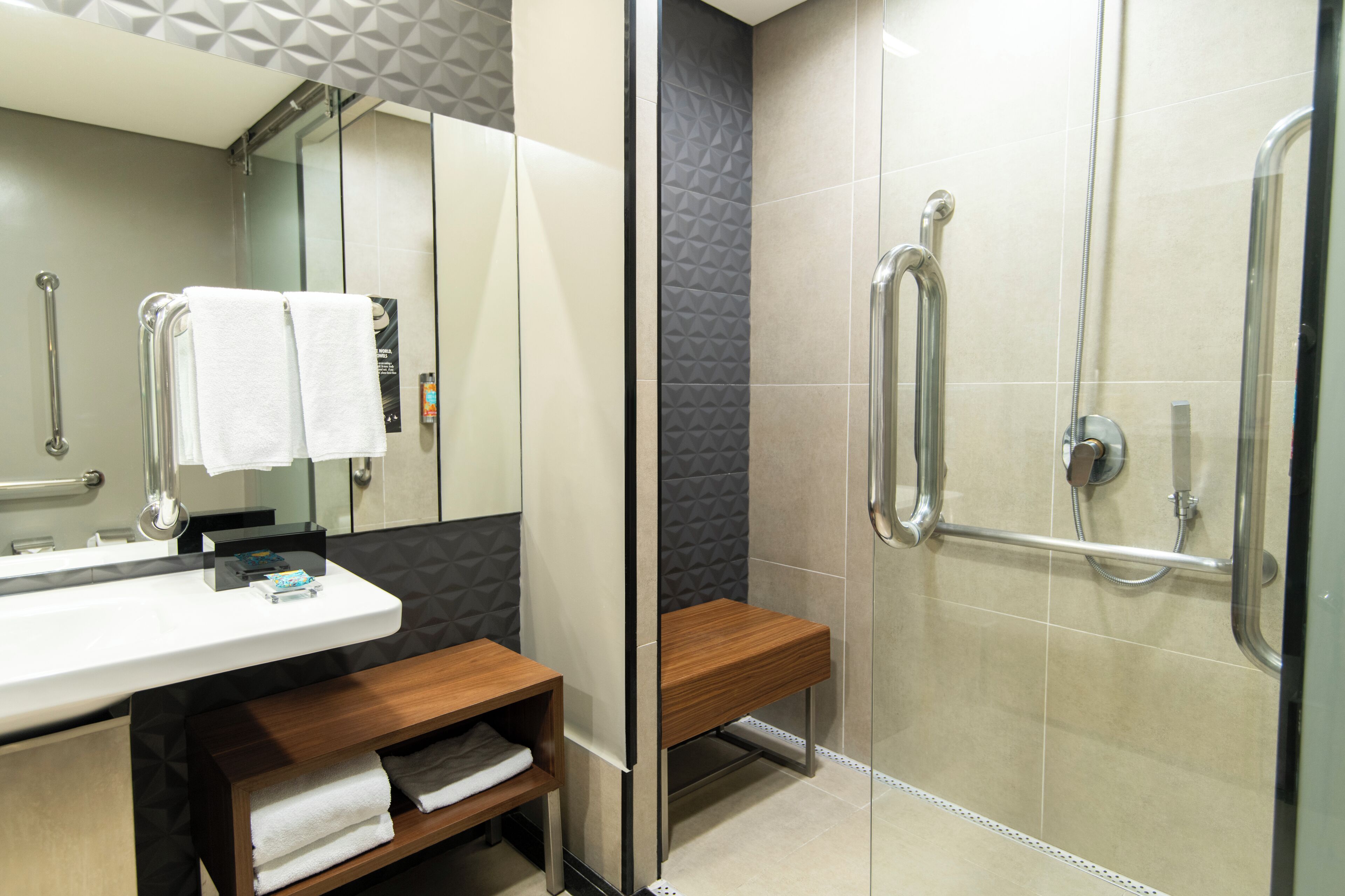 Un baño moderno con cabina de ducha de vidrio, un banco de madera y un mueble de tocador con espejo y toallero.