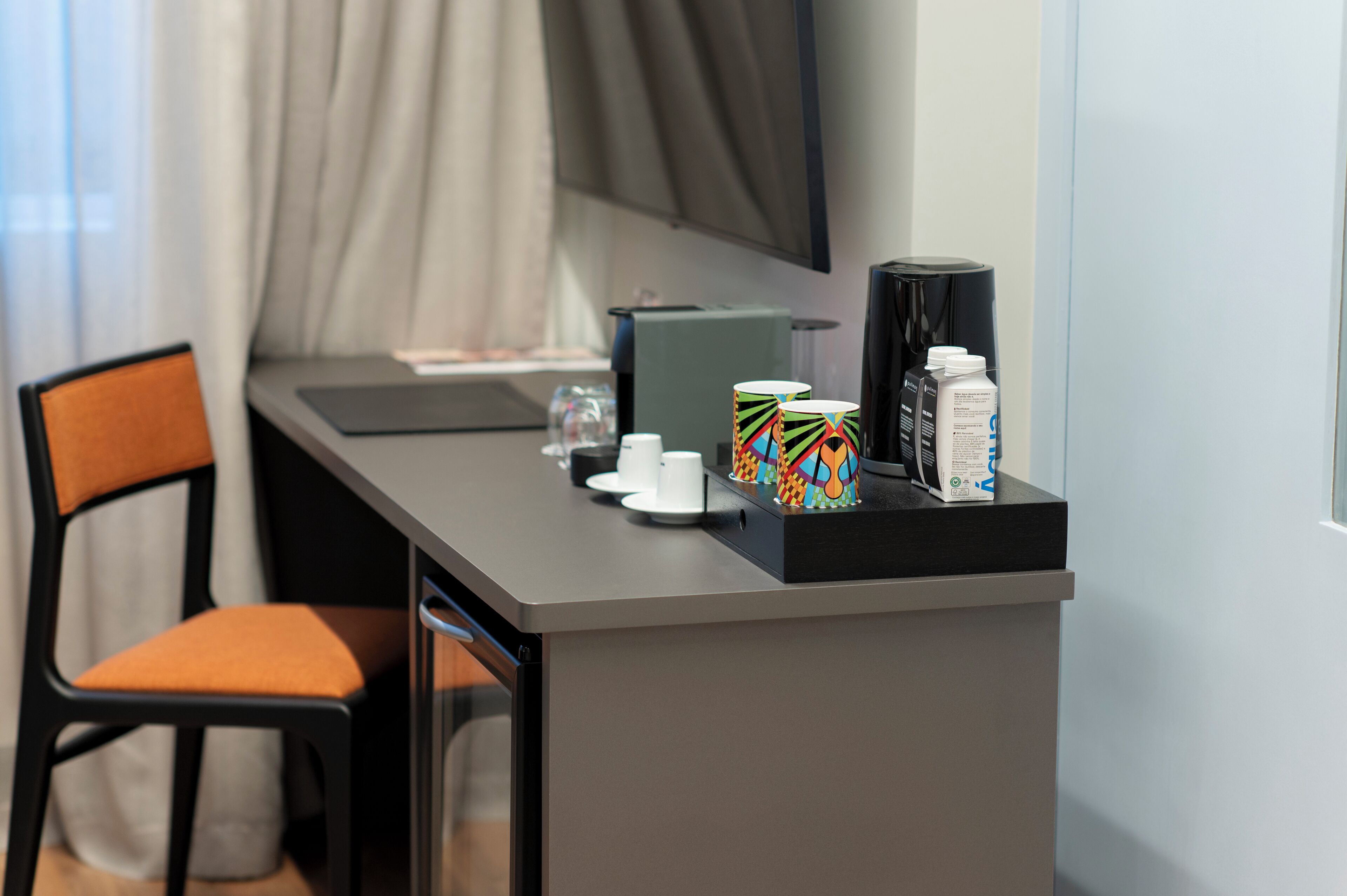 Quarto de hotel moderno com escrivaninha, cadeira, máquina de café, caneca, copo e caixa de lenços.