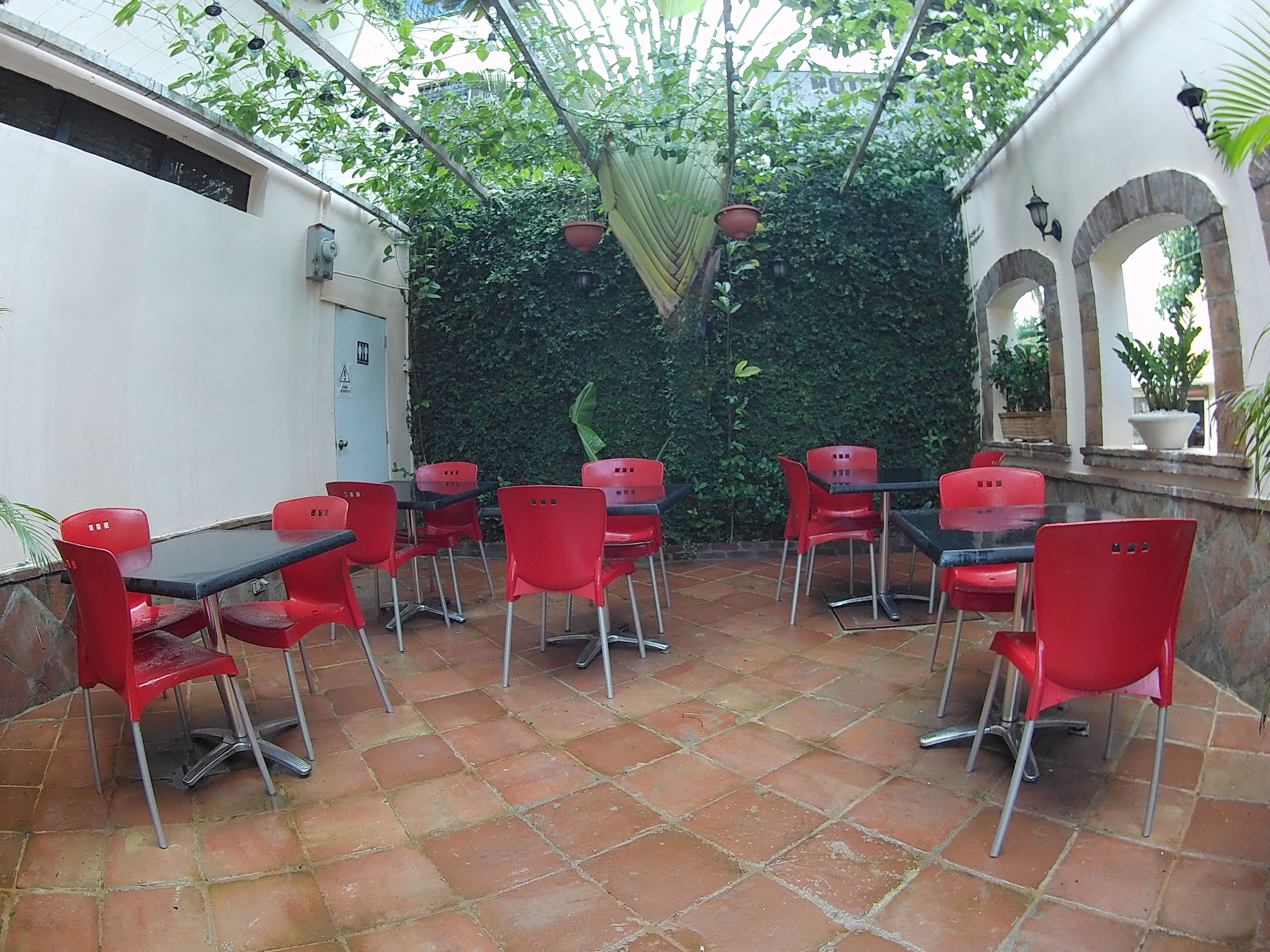 Une terrasse avec des chaises et des tables rouges, entourée d’une haie verte et de plantes en pot.