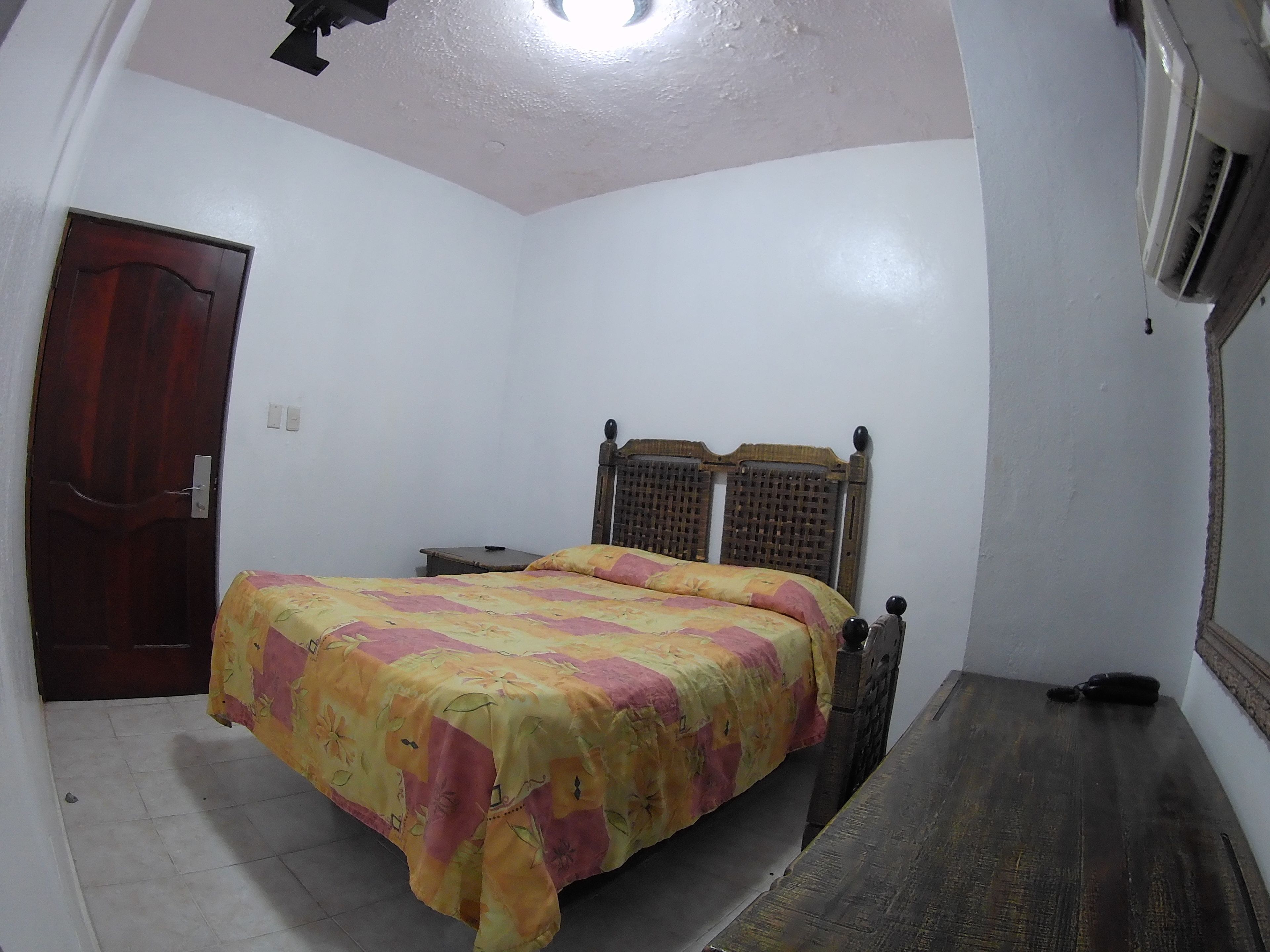 A bedroom with a bed, a wooden headboard, a nightstand, a door, and a wall-mounted air conditioning unit.
