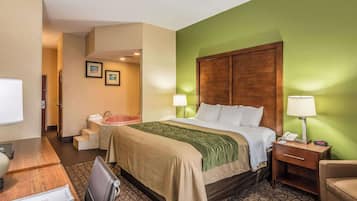 Habitación de hotel con cama, escritorio, silla, mesita de noche, lámpara y baño con lavamanos y espejo.