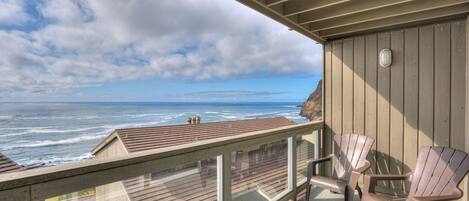 A balcony with a view of the ocean, wooden deck, and Adirondack chairs.