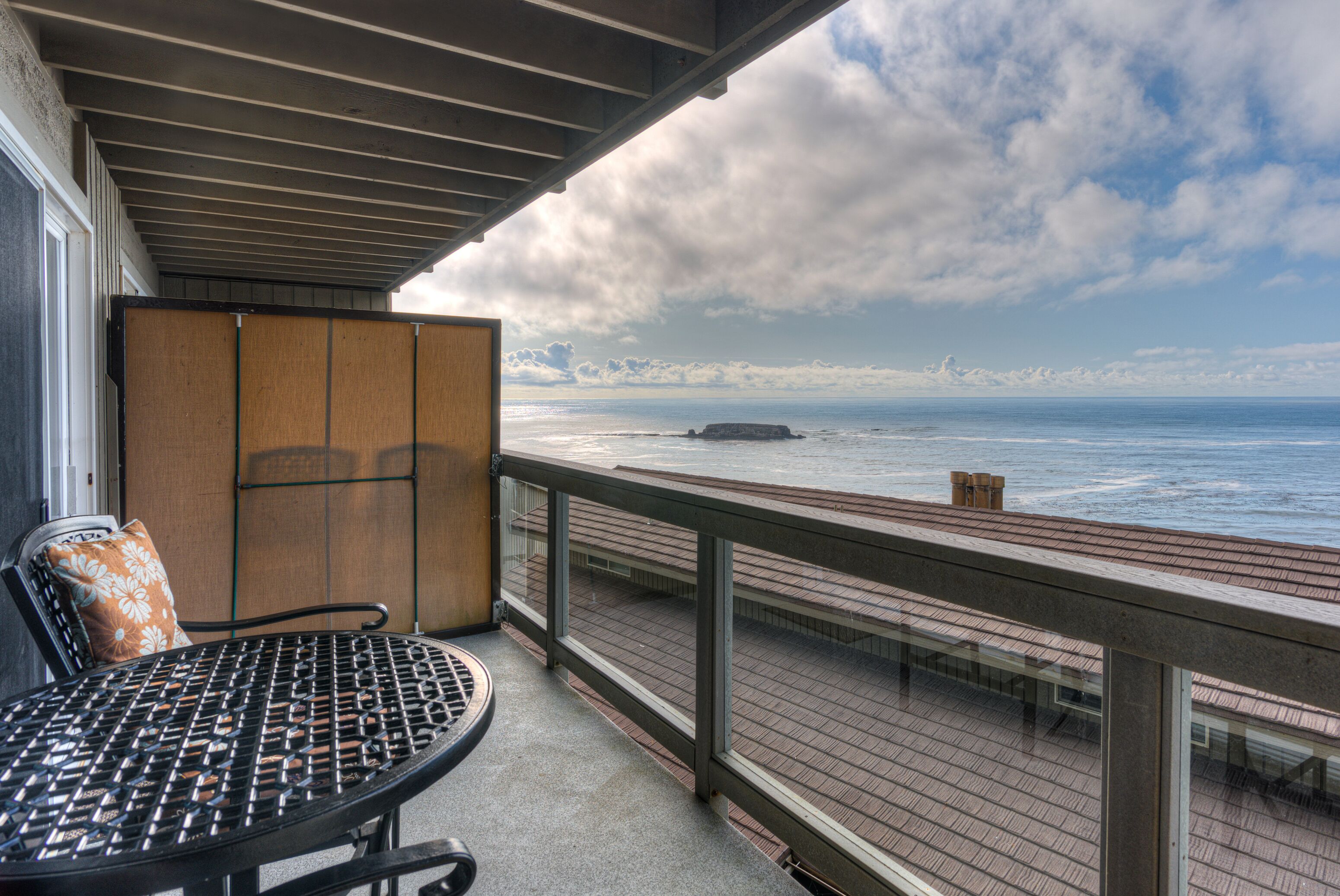 A balcony with a metal table and chairs, overlooking a wooden deck and the ocean.