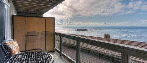 A balcony with a metal table and chairs, overlooking a wooden deck and the ocean.