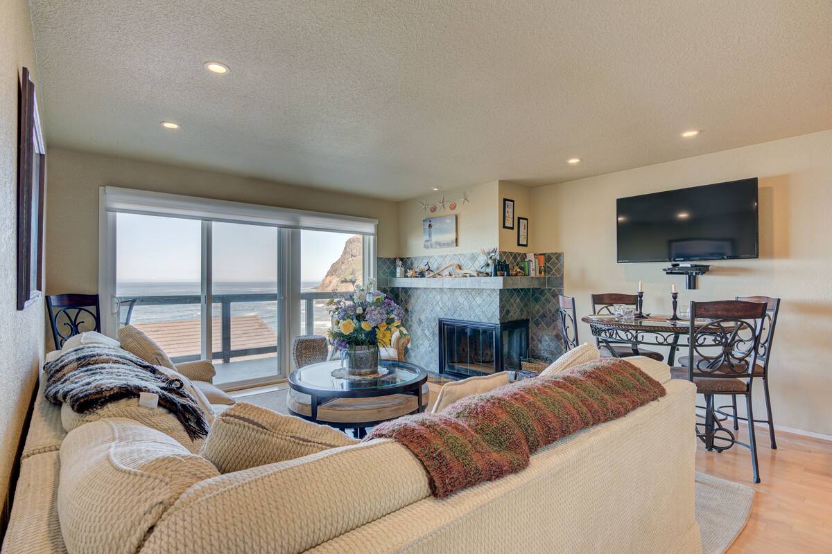 A spacious living room with a large sofa, a fireplace, a dining area, and a balcony with a view.
