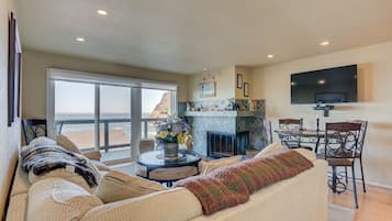 A spacious living room with a large sofa, a fireplace, a dining area, and a balcony with a view.