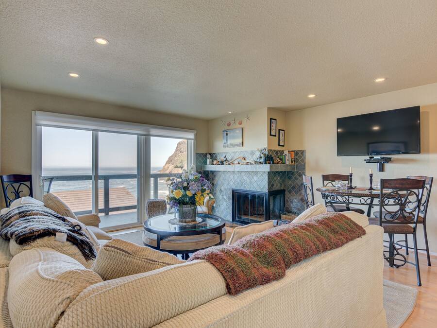A spacious living room with a large sofa, a fireplace, a dining area, and a balcony with a view.