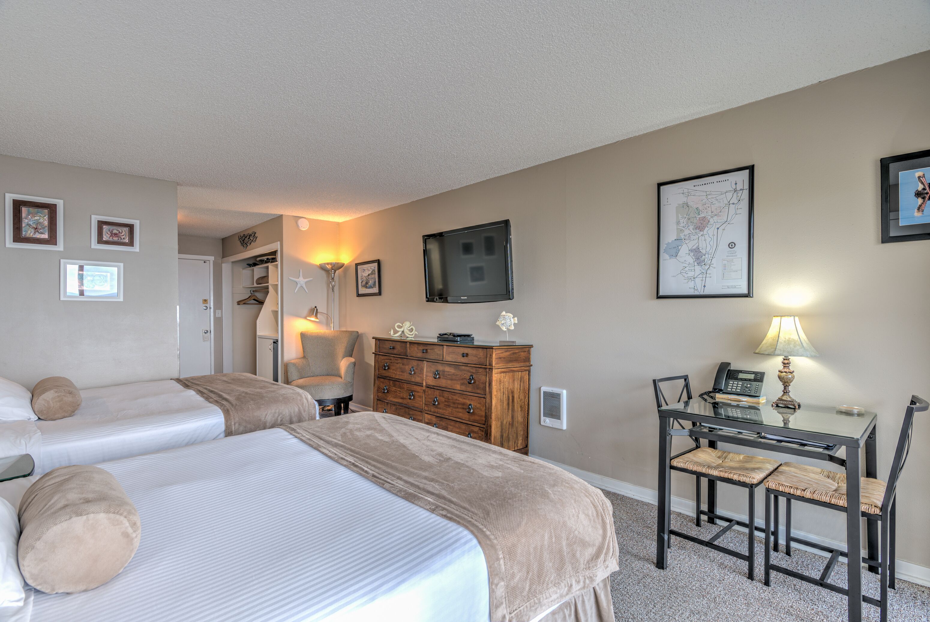 A hotel room with two beds, a desk with a chair, a dresser, a TV mounted on the wall, and framed pictures.