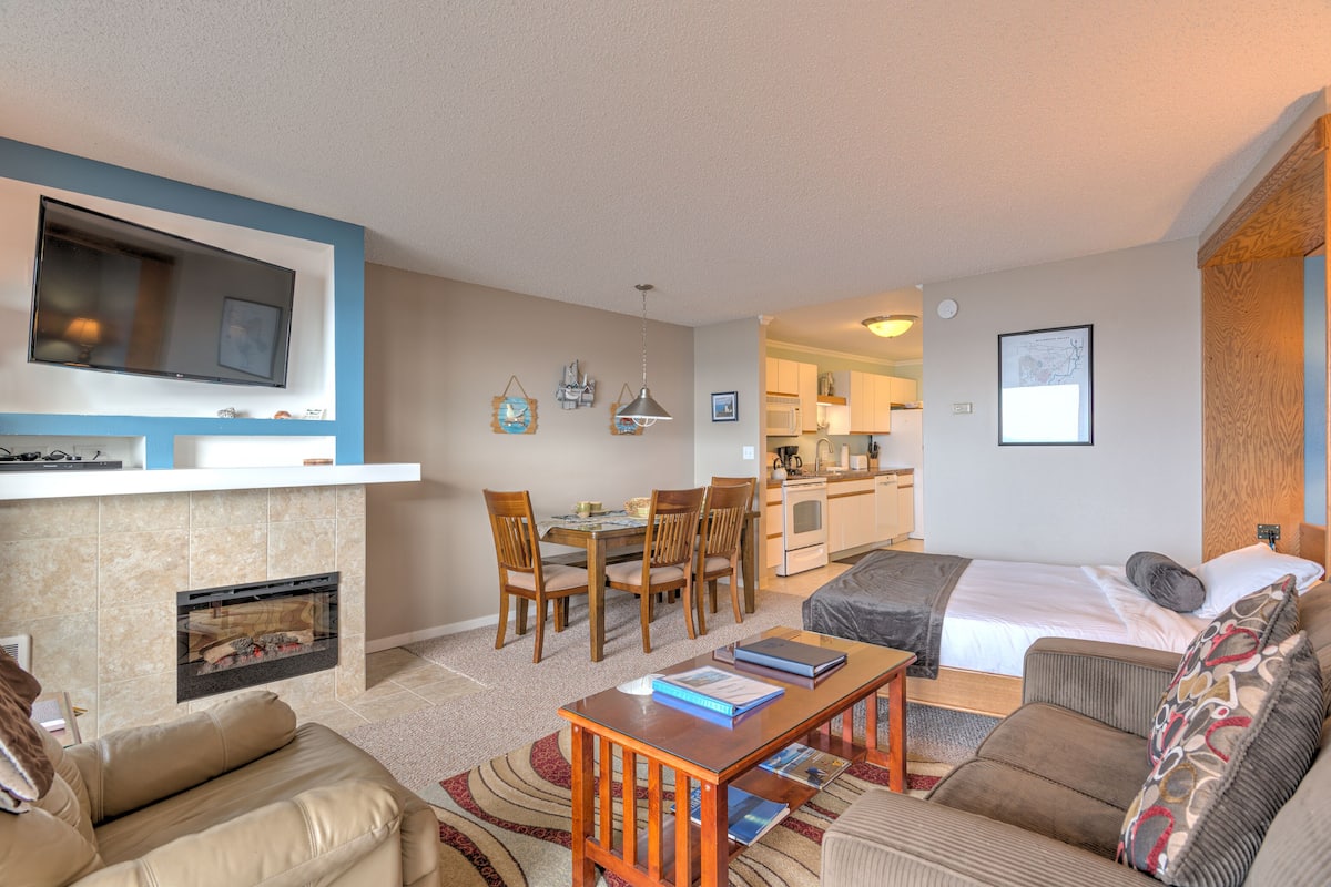 A modern living room with a fireplace, a dining area, a kitchen, and a bedroom.