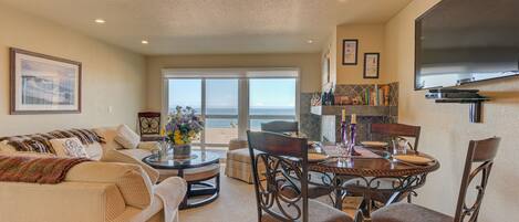 A living room with a sofa, coffee table, and dining table with chairs. There is a large window with a view of the ocean.