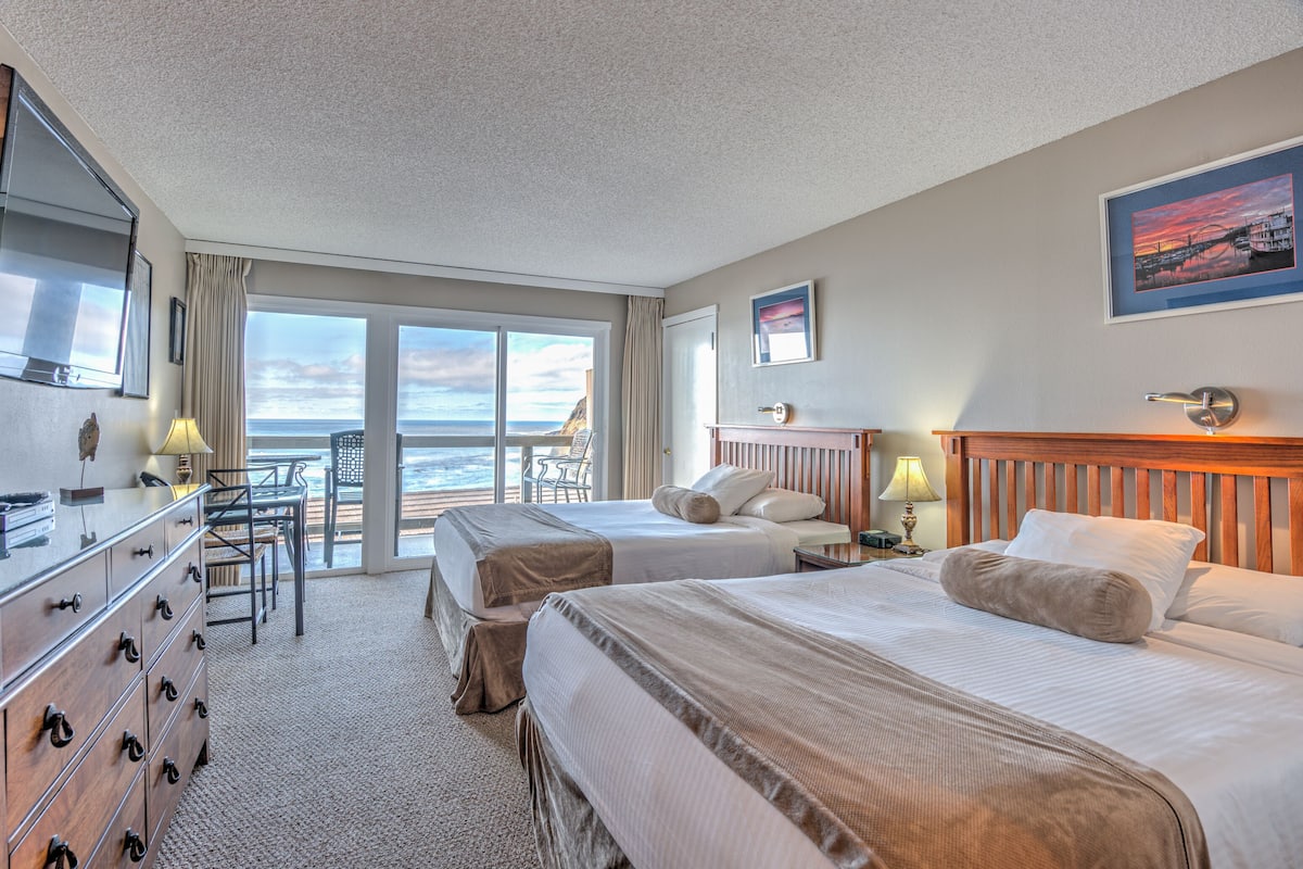 A hotel room with two beds, a TV, a desk with a chair, and a view of the ocean.