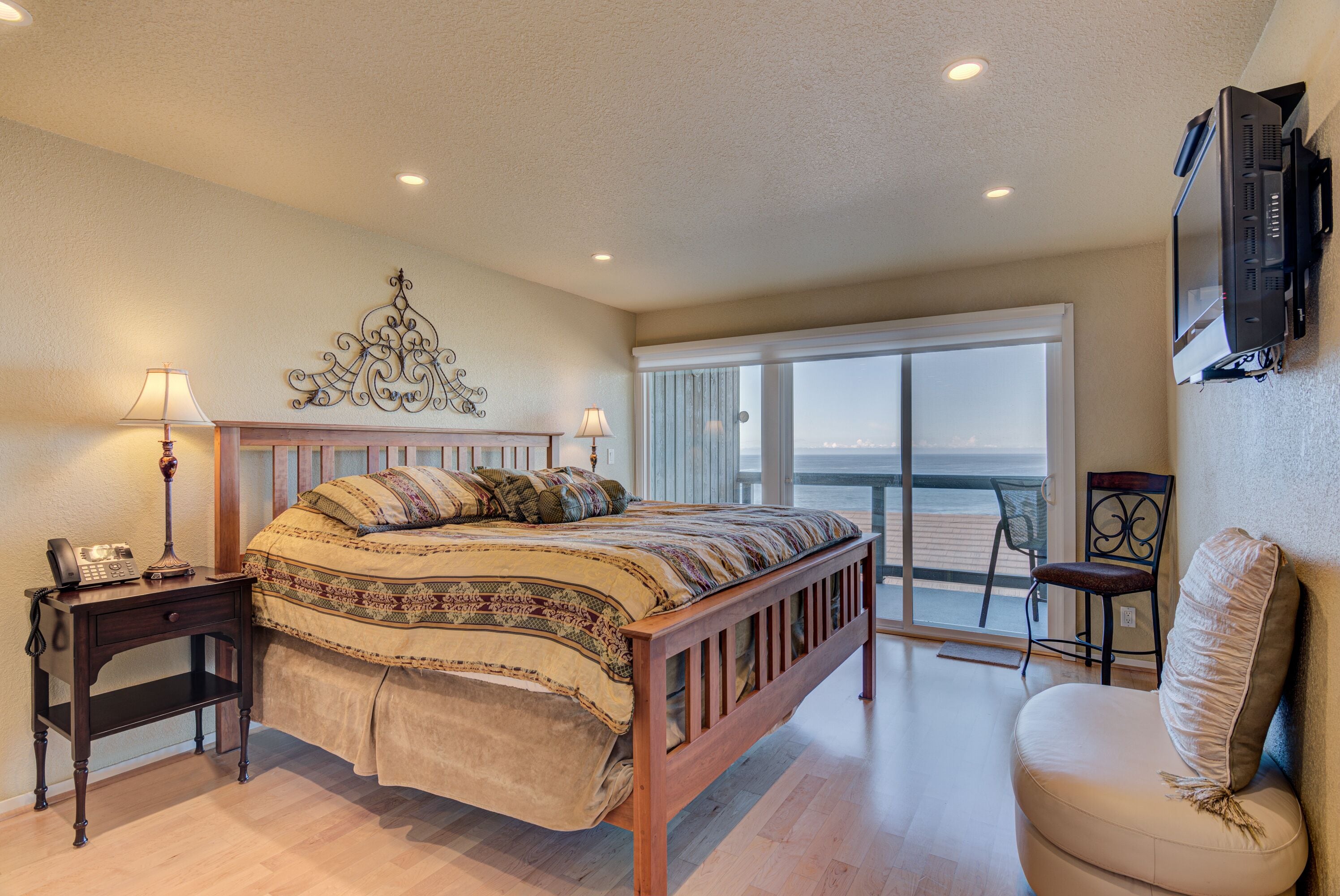 A bedroom with a large bed, a nightstand with a lamp, a chair, and a view of the ocean through a sliding glass door.