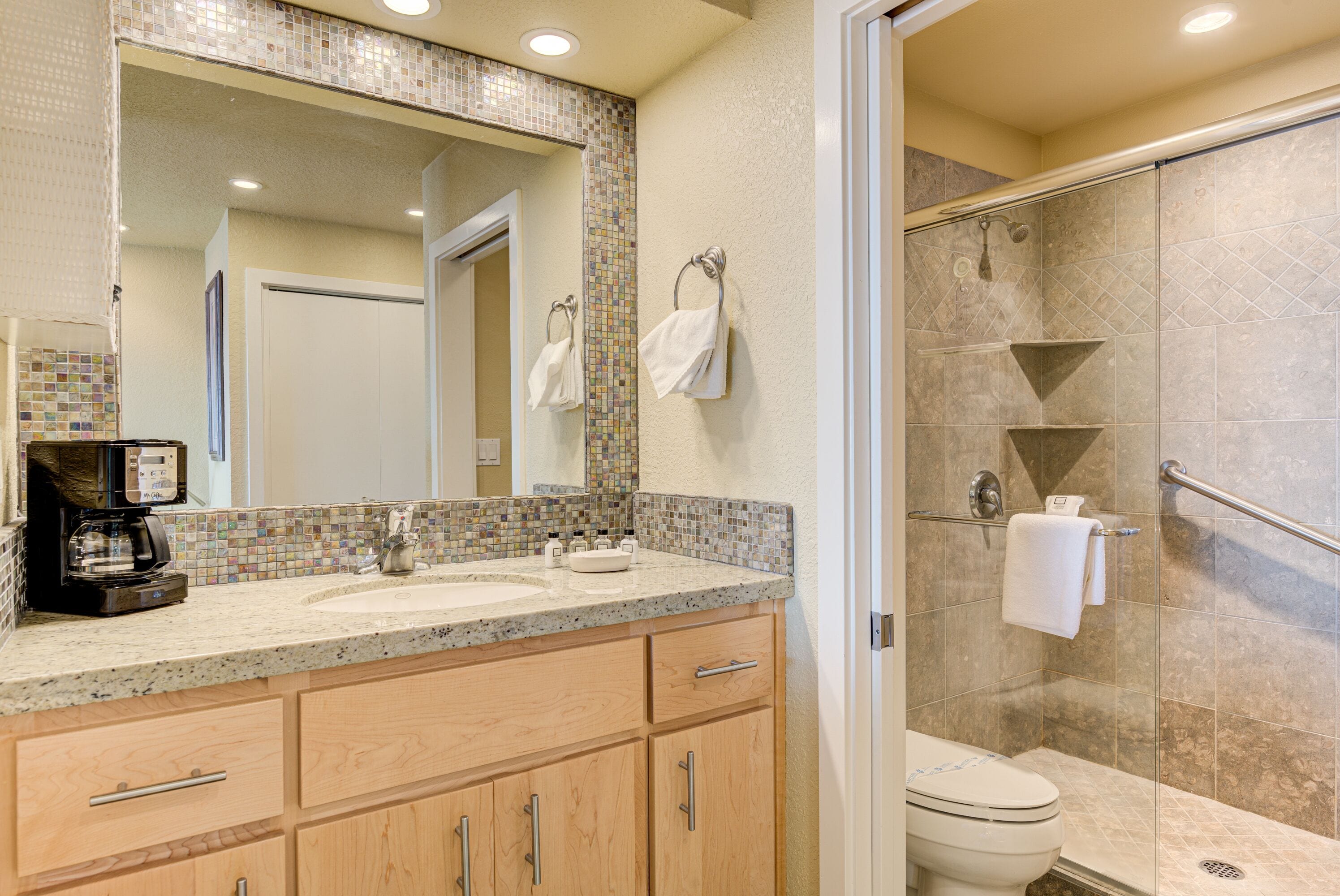 Un baño con lavamanos doble, cafetera, ducha con puerta de cristal y salpicadero de azulejos.