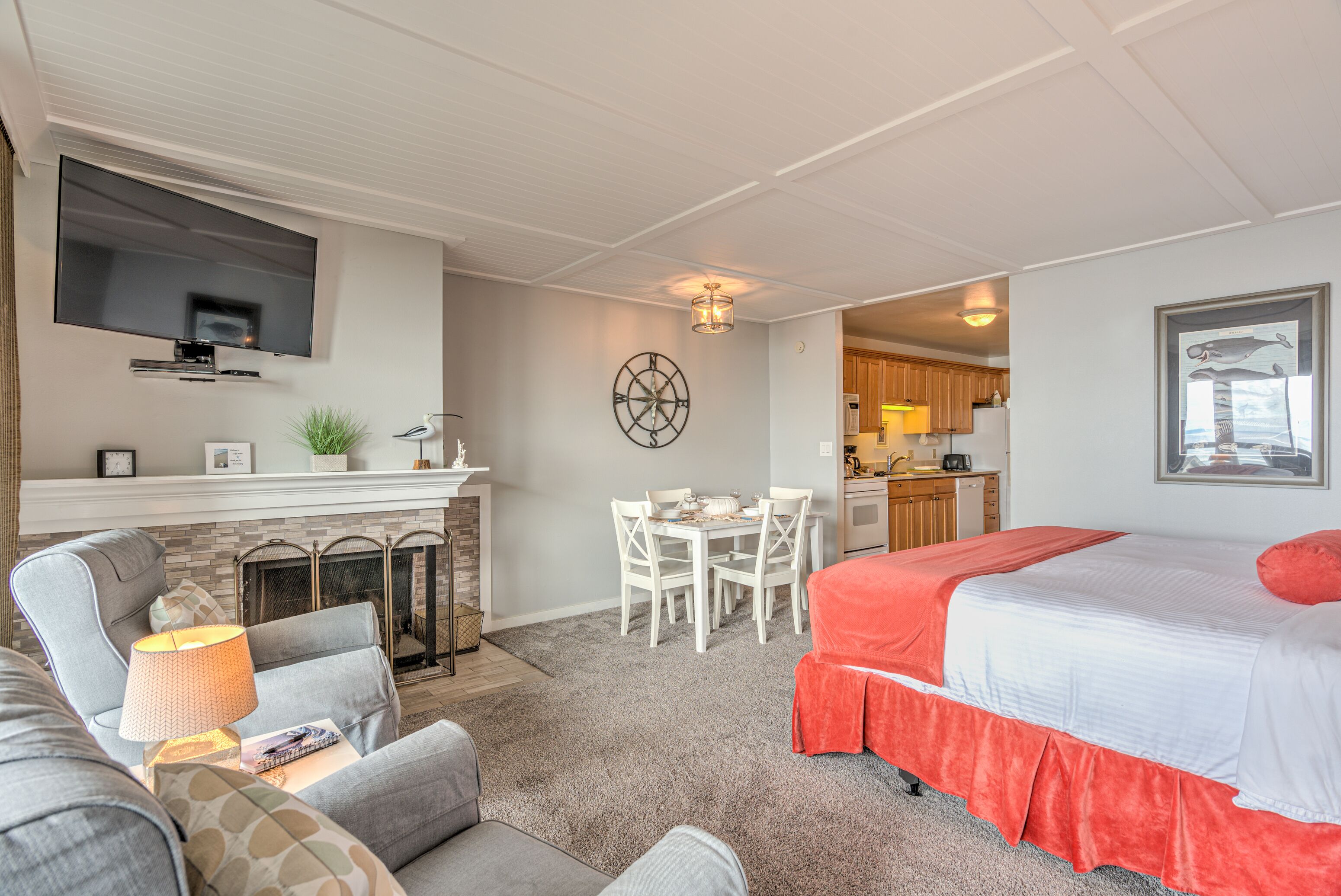 A hotel room with a flat-screen TV mounted above a fireplace, a dining area with a table and chairs, and a bed with a red accent.