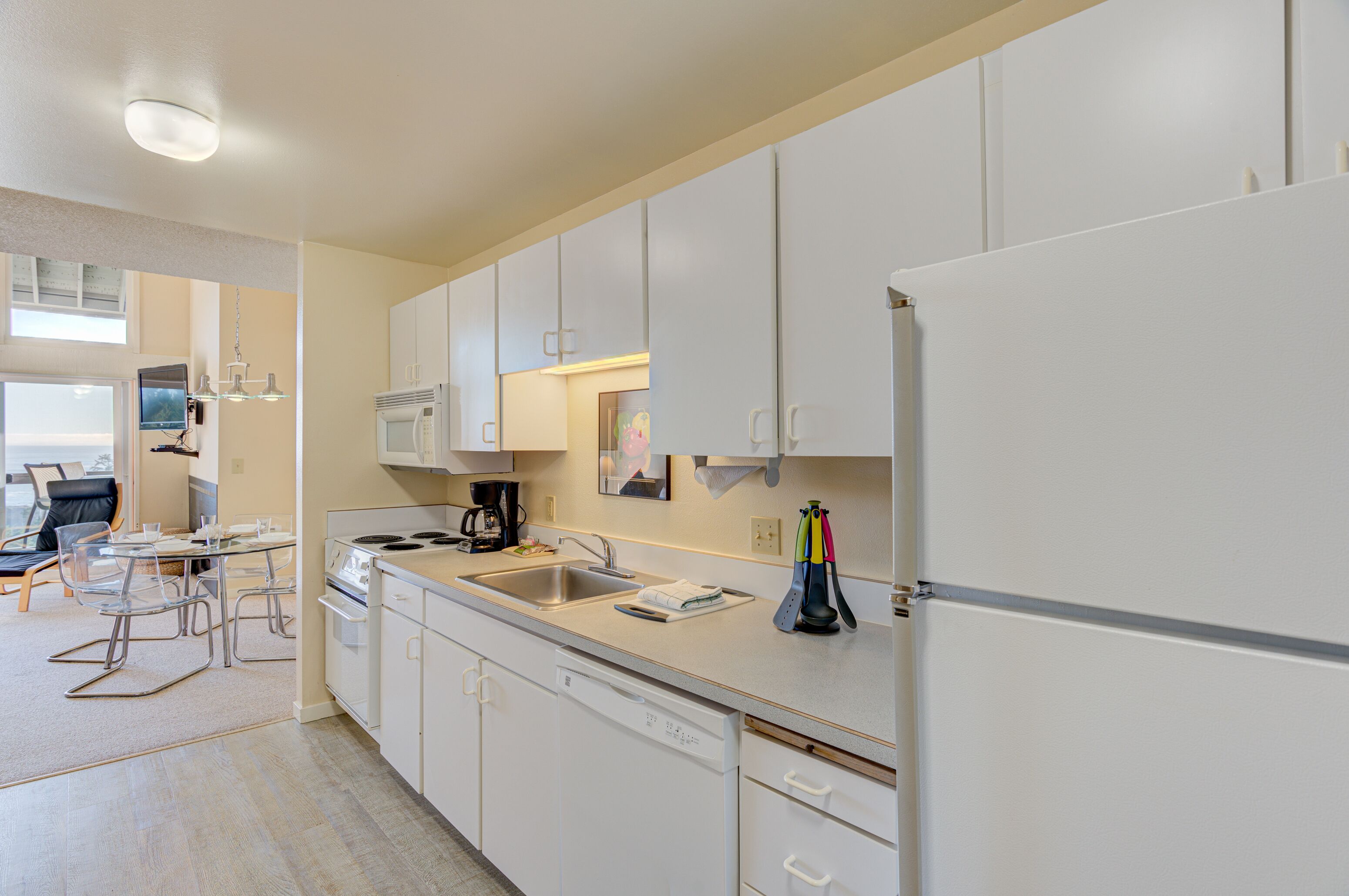 Una cocina moderna con armarios blancos, un lavamanos de acero inoxidable y un lavavajillas. Hay una mesa de comedor con sillas y una cafetera en la encimera.