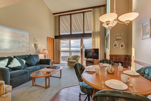 A living room with a sofa, coffee table, and a dining area with a table set for four. There is a television and a bookshelf in the background.