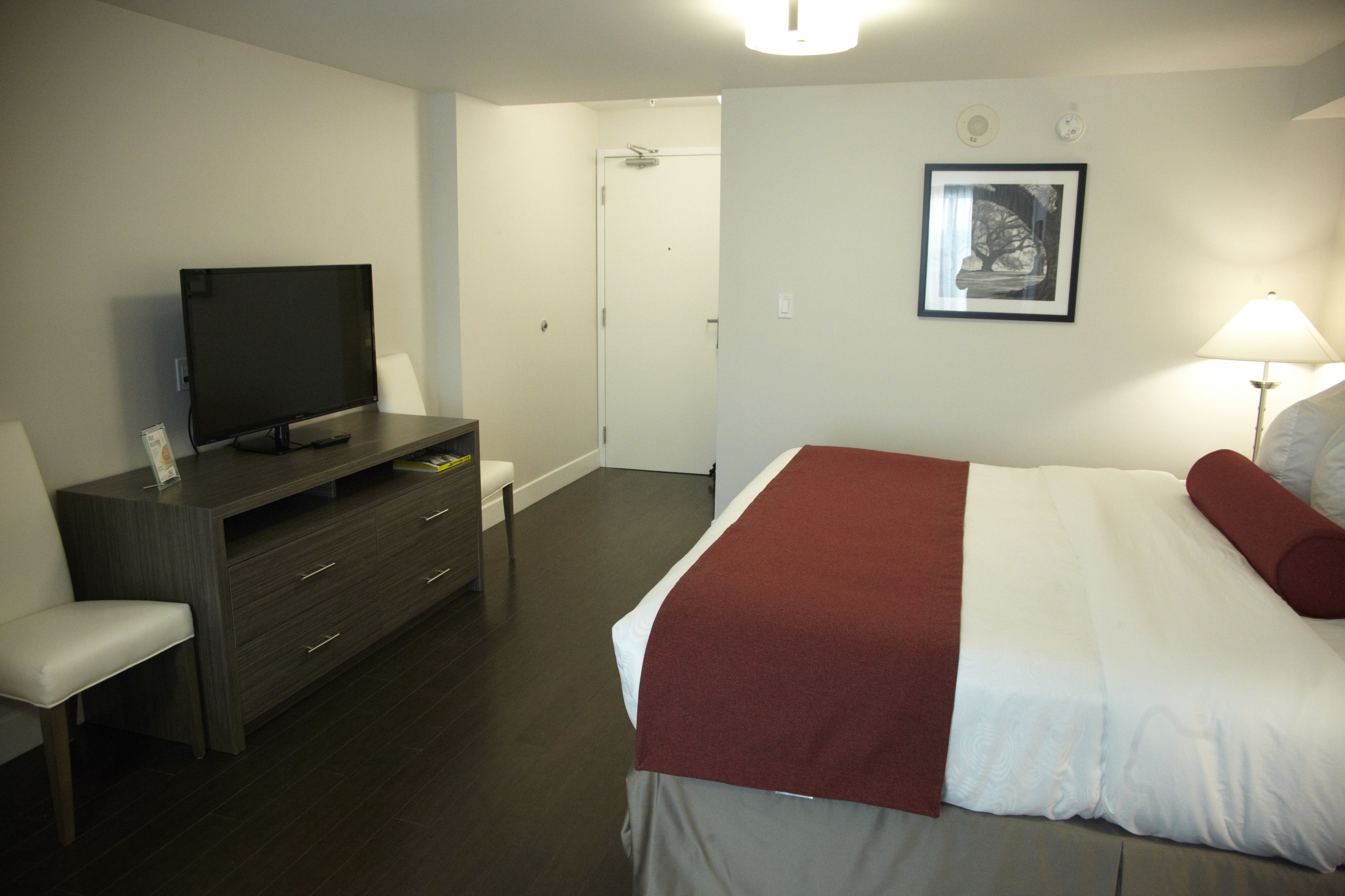 A hotel room with a bed, a TV on a stand, a desk with drawers, a chair, and a door leading to a bathroom.