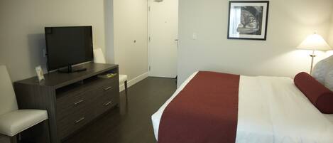 A hotel room with a bed, a TV on a stand, a desk with drawers, a chair, and a door leading to a bathroom.