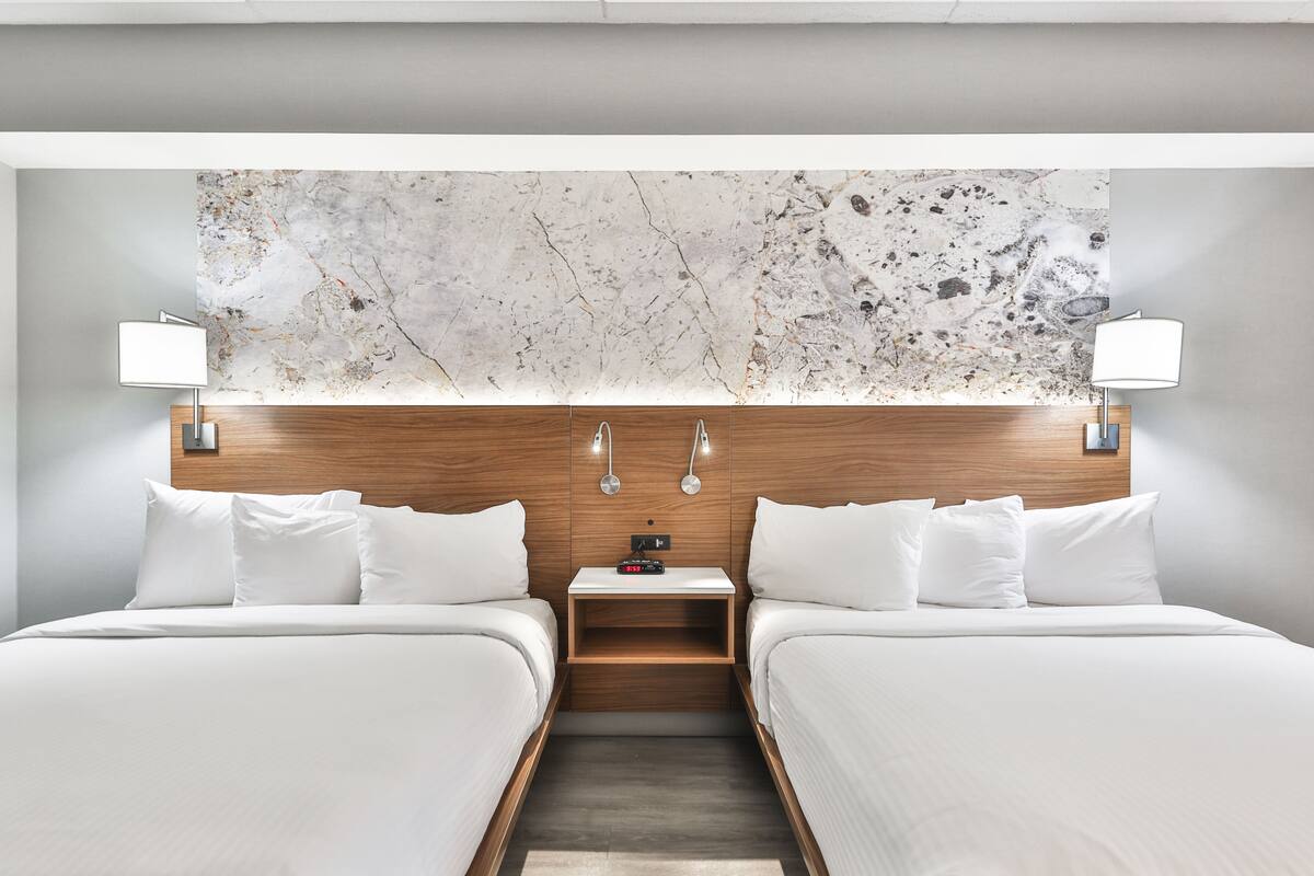 Habitación de hotel con dos camas, cabecero de madera, pared de mármol y dos lámparas de noche.
