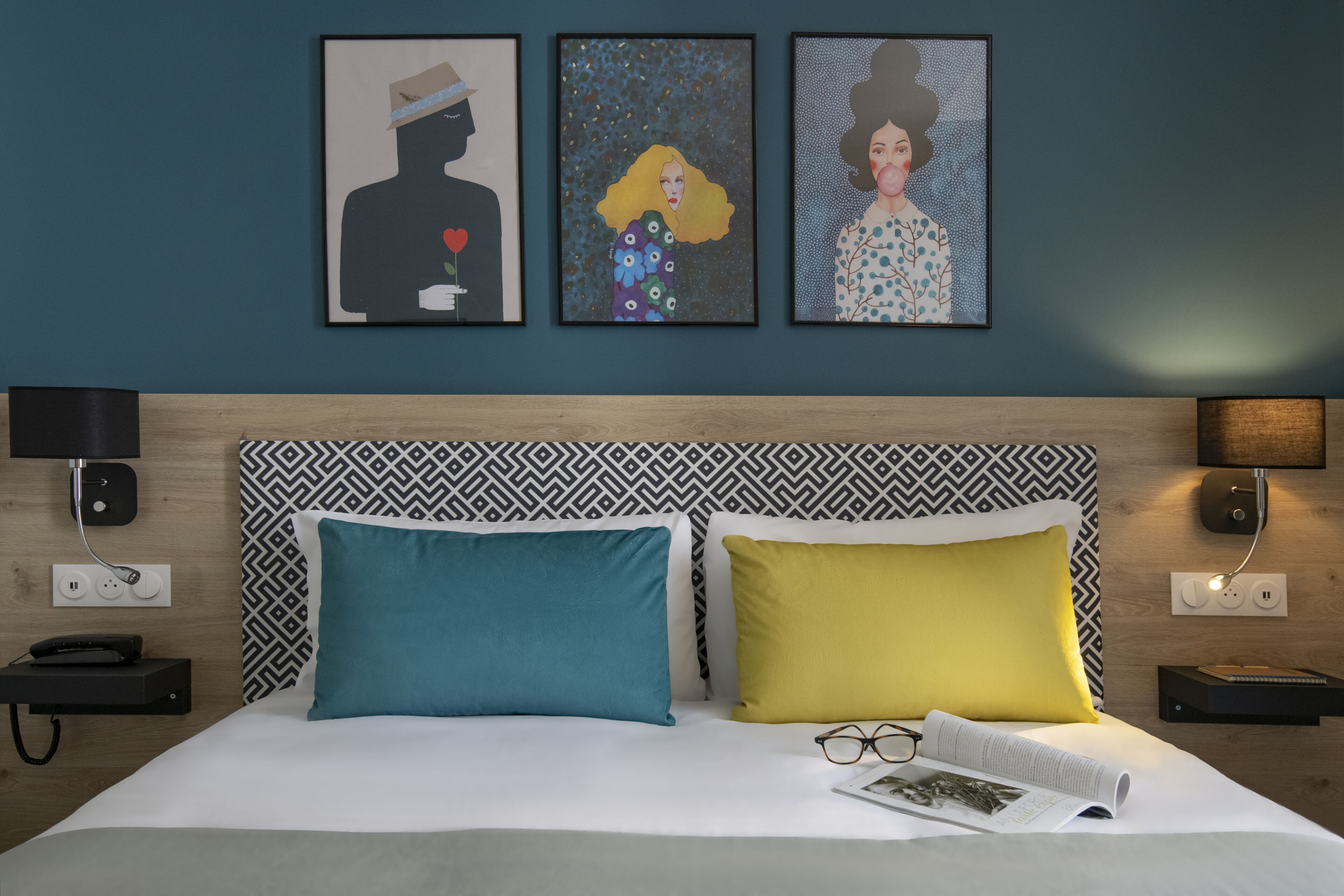 Une chambre d’hôtel avec un lit, deux oreillers, une table de chevet, une lampe, un téléphone et trois œuvres d’art encadrées accrochées au mur.