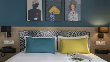 Une chambre d’hôtel avec un lit, deux oreillers, une table de chevet, une lampe, un téléphone et trois œuvres d’art encadrées accrochées au mur.