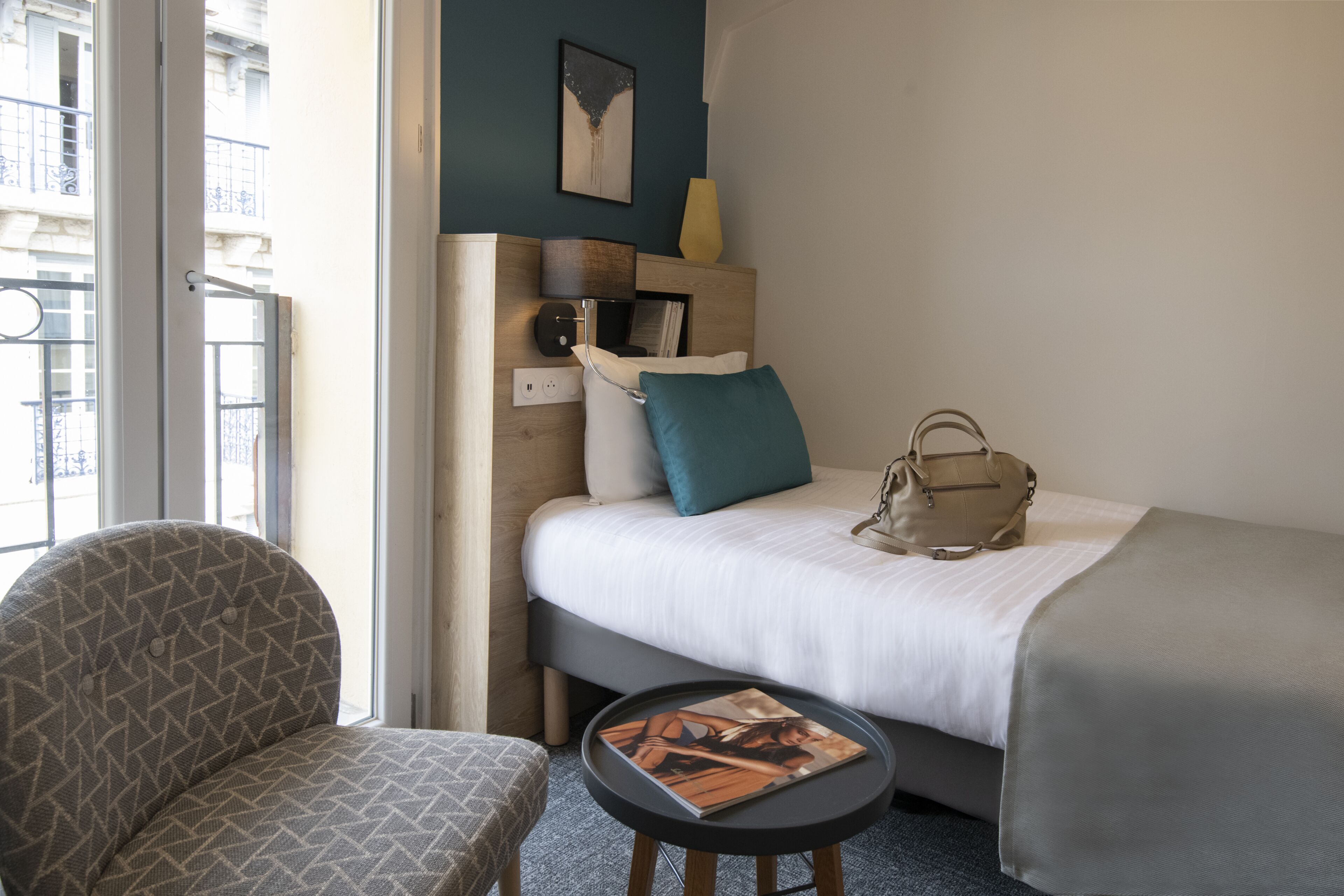 A hotel room with a bed, a chair, a small table, and a window with a view of buildings.