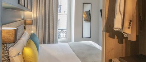 A hotel room with a bed, a bedside table, a lamp, a window with curtains, a wardrobe, and a suitcase.