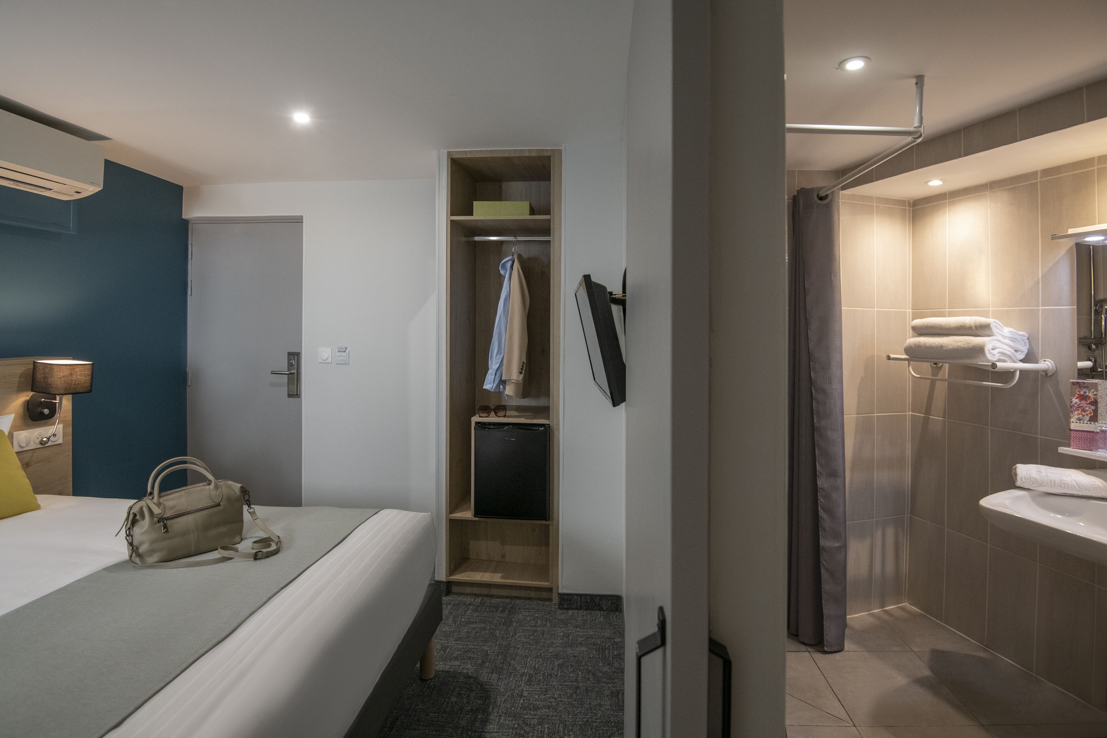 Habitación de hotel con cama, mesita de noche con lámpara, armario, refrigerador pequeño y baño con lavamanos y espejo.