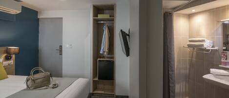 Habitación de hotel con cama, mesita de noche con lámpara, armario, refrigerador pequeño y baño con lavamanos y espejo.