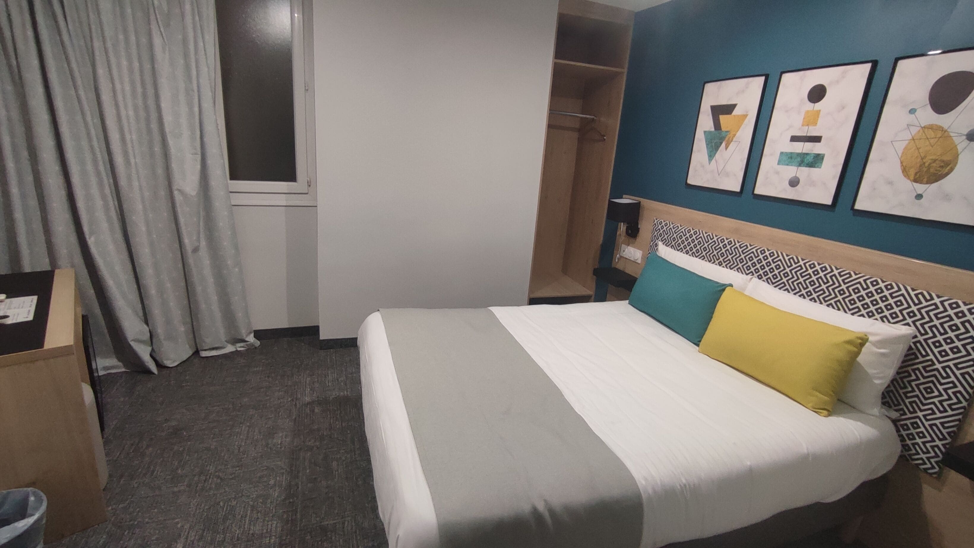 Une chambre d’hôtel avec un lit, une armoire, une fenêtre avec des rideaux et des œuvres d’art murales.